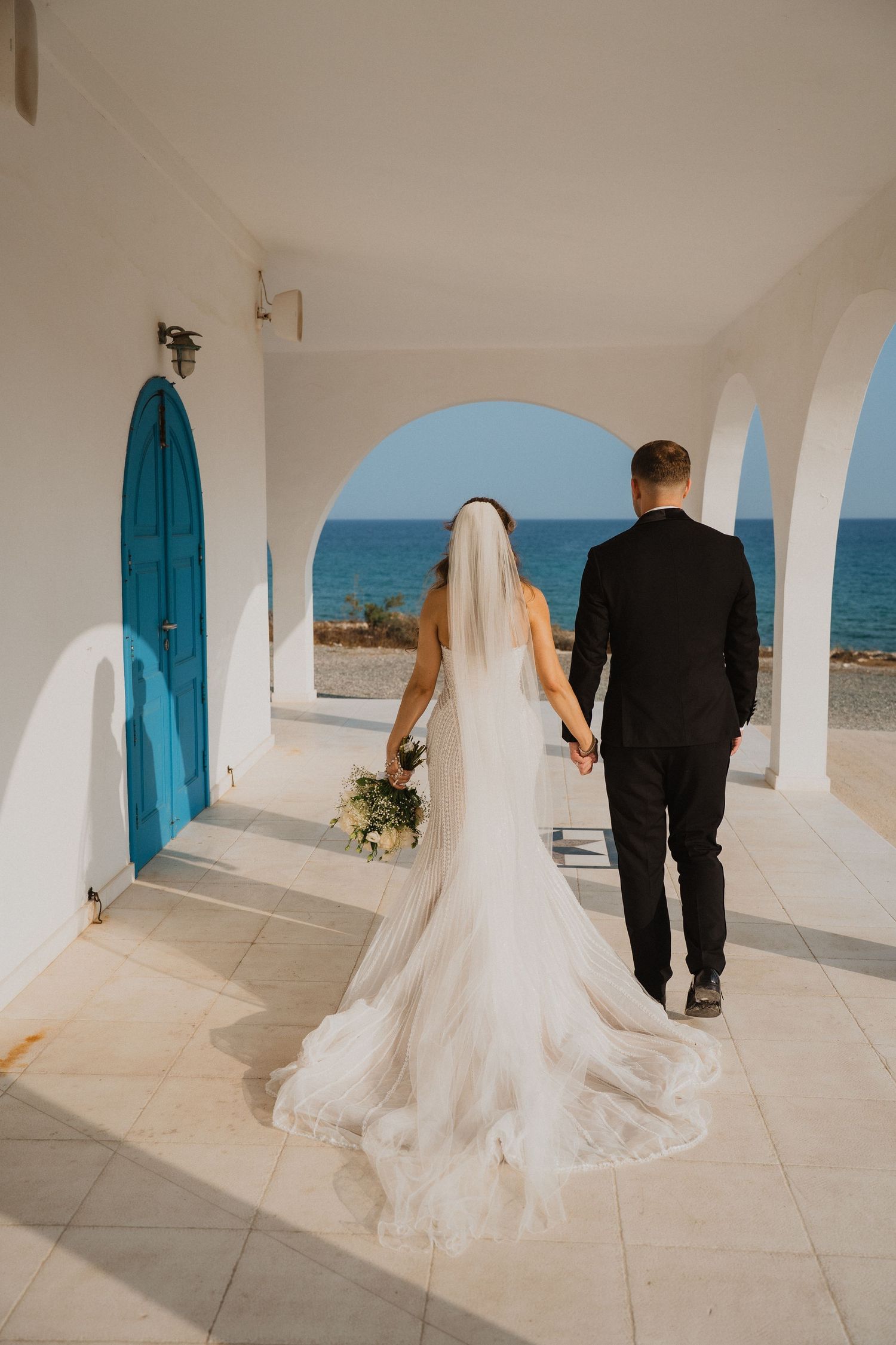 Bride and groom walk toward blue Mediterranean sea through white arched chapel doorway, veil trailing behind.