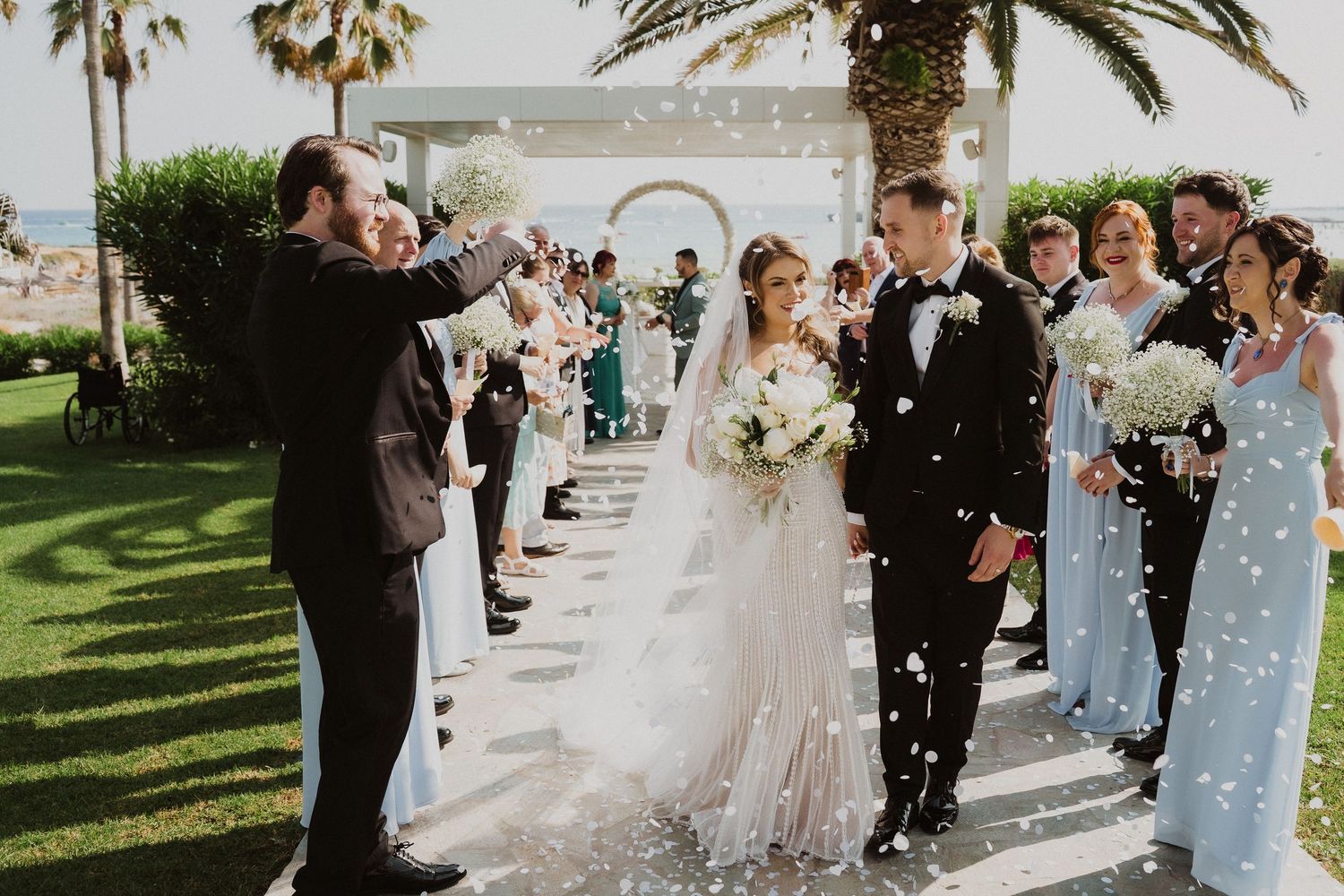 Bride and groom walk down aisle as guests toss white confetti at outdoor wedding ceremony with palm trees.