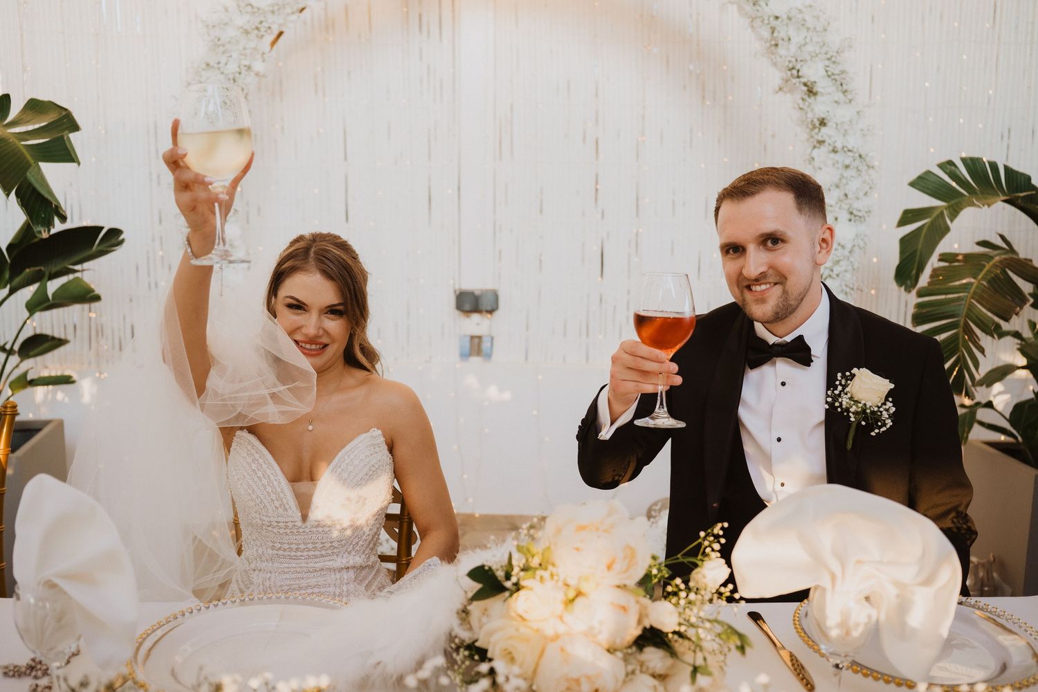 Bride and groom raise glasses in a toast at their sweetheart table decorated with white floral arrangements.