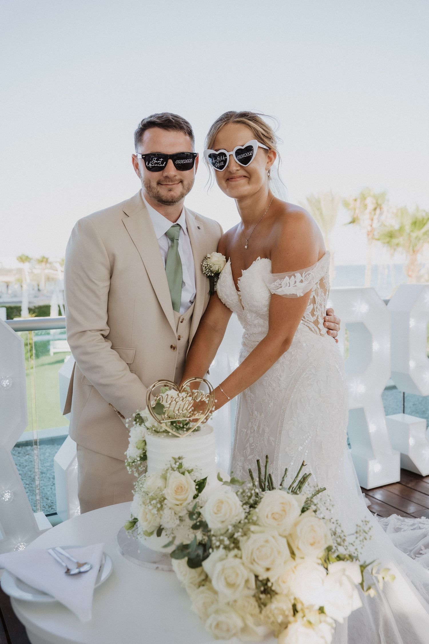 Wedding couple in sunglasses pose with elegant white floral bouquet at outdoor reception venue.