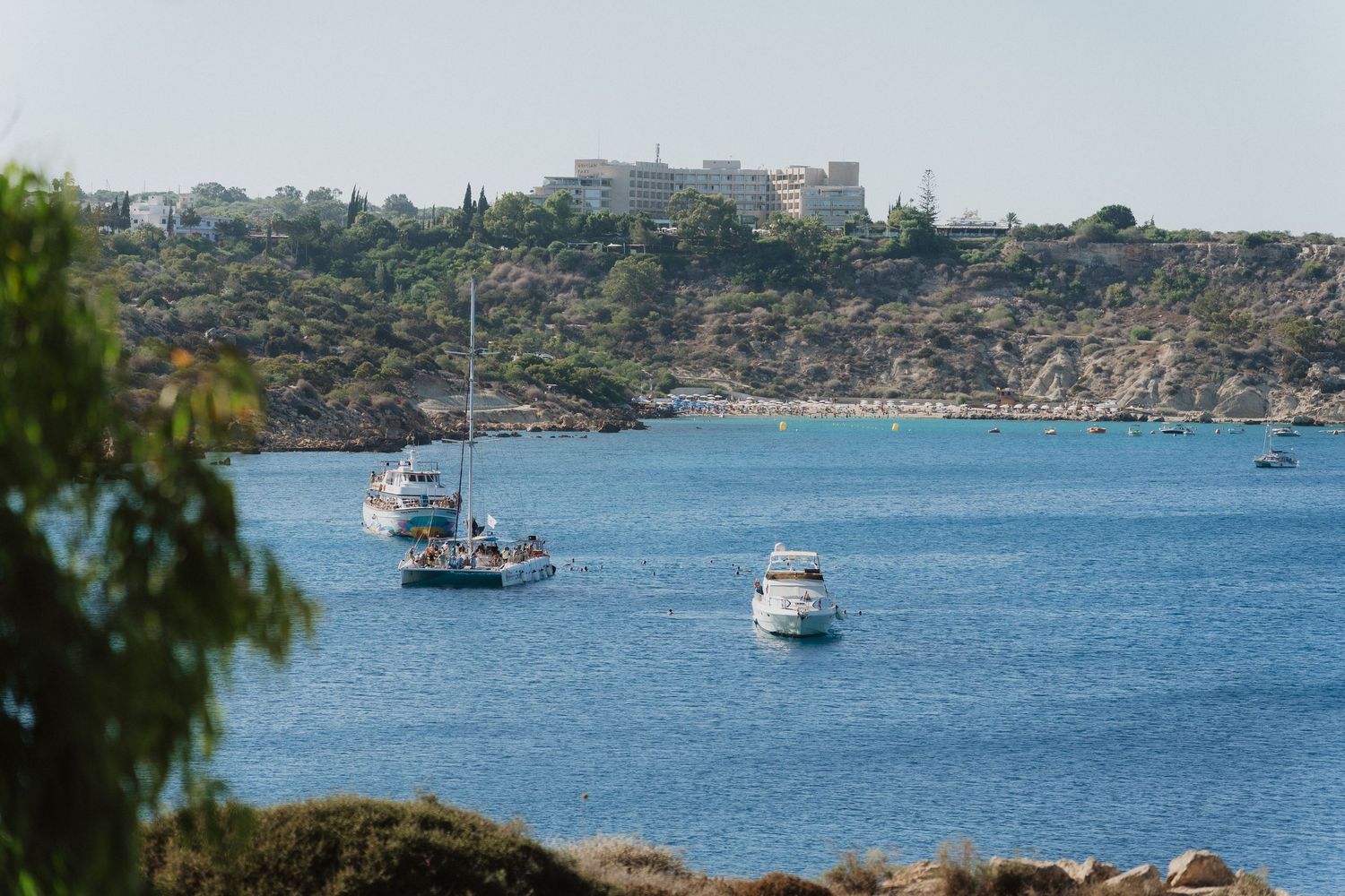 Scenic Mediterranean coastline with boats anchored in blue waters and buildings dotting the hillside.