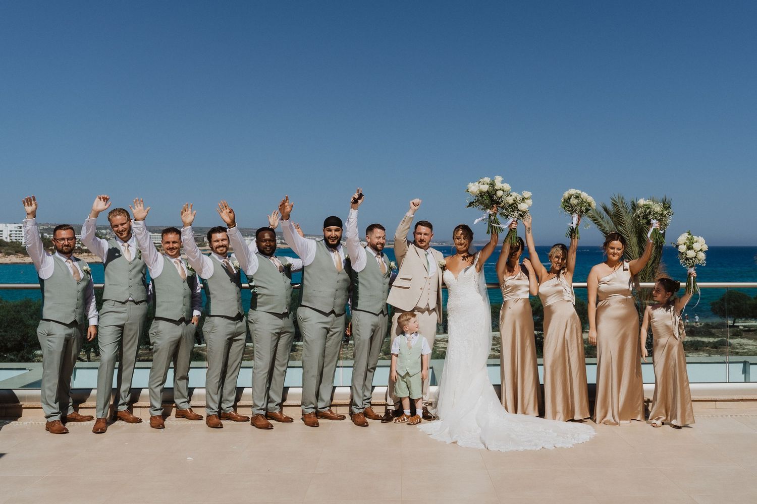 Wedding party poses playfully on a sunny beach with bridesmaids in beige dresses and groomsmen in gray suits.