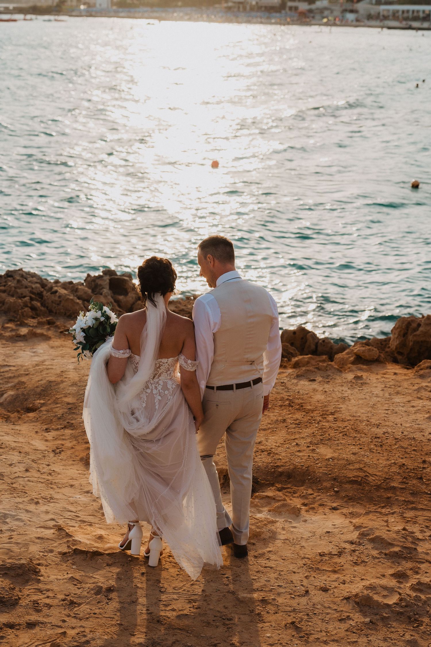 A couple in wedding attire walks along a rocky coastal cliff during sunset, with ocean waves visible in the background.