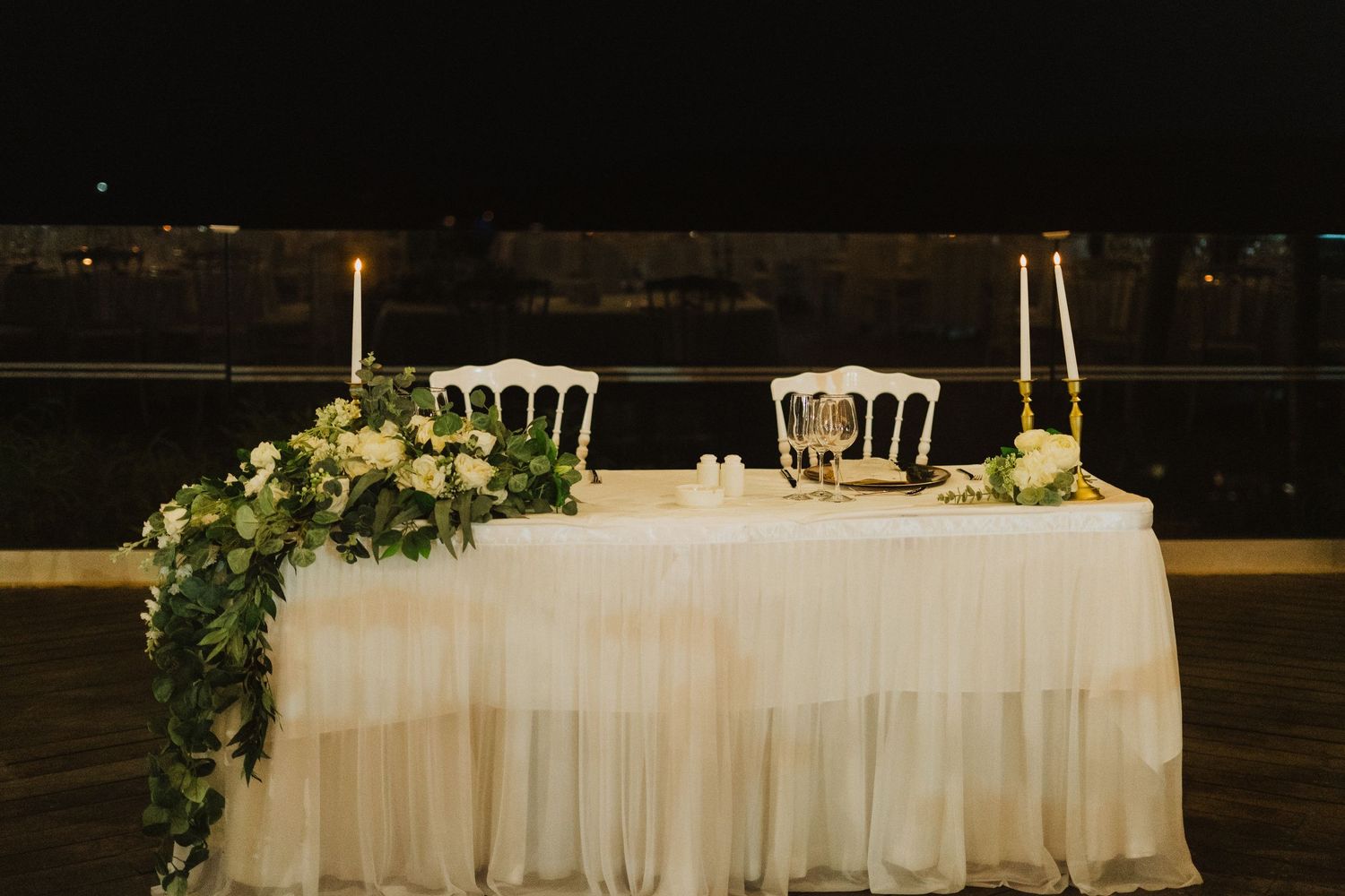 Wedding reception sweetheart table decorated with white linens and cascading greenery at night.