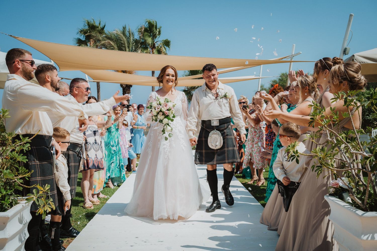 Guests throw confetti at newlyweds walking down outdoor aisle under beige canopy.