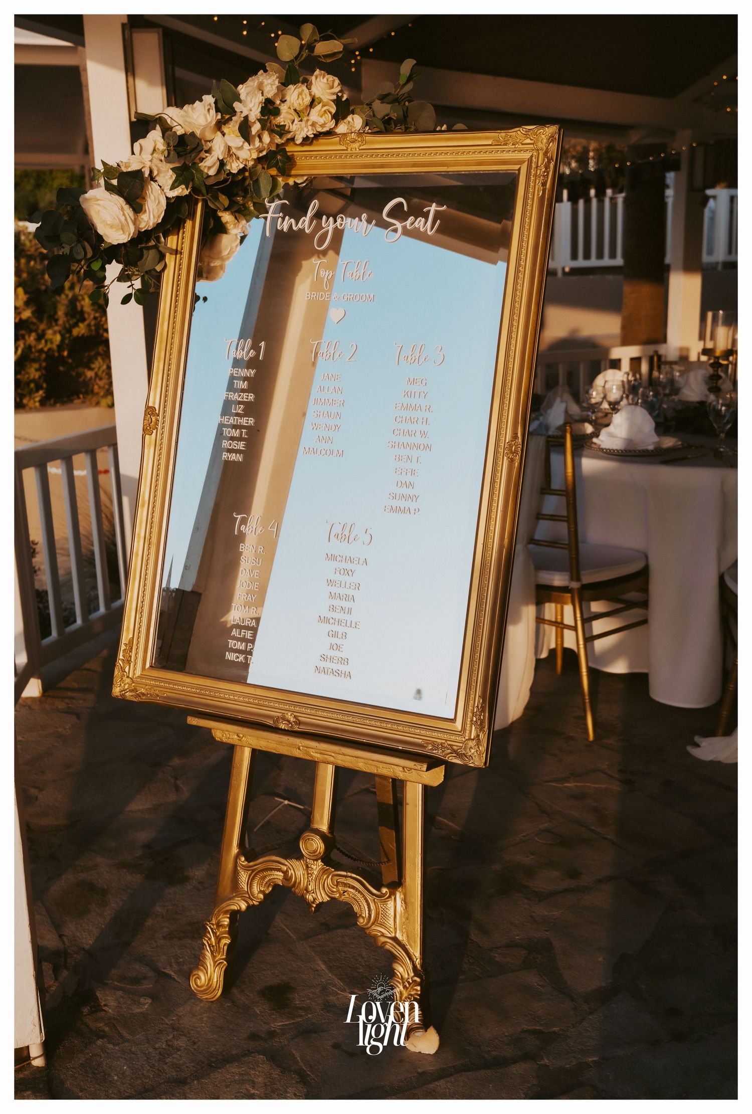 Ornate gold frame on easel displaying wedding seating chart with floral accents.