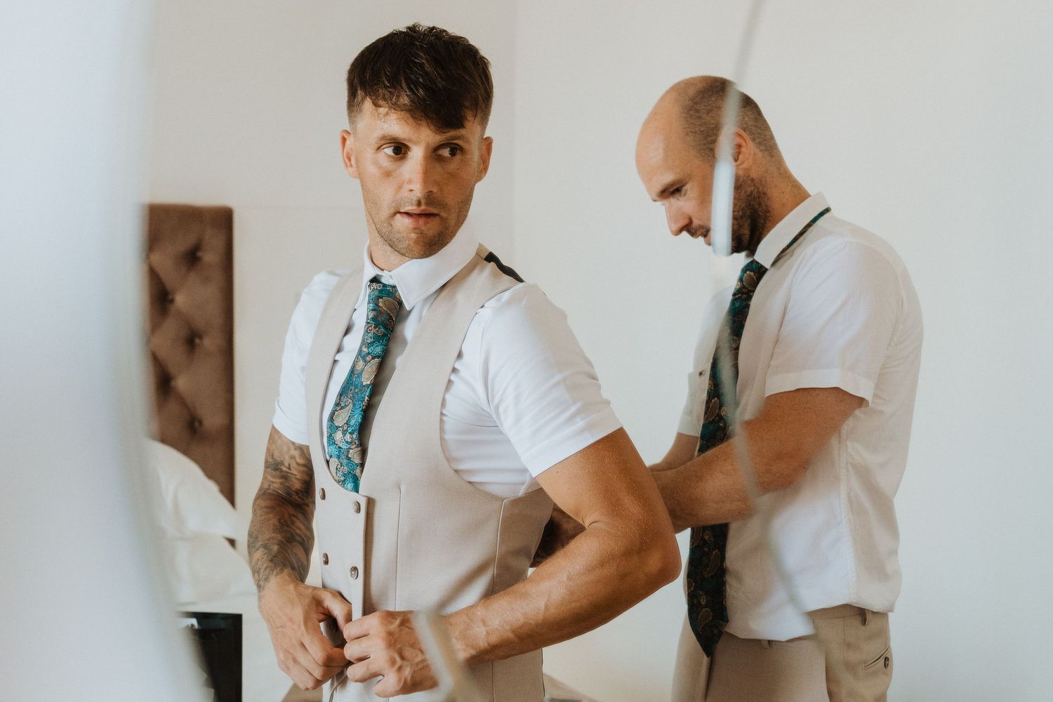 Two people in white dress shirts and suspenders getting ready for a formal event with one adjusting their outfit.