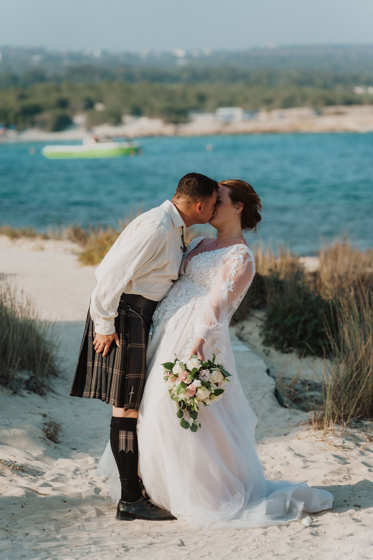 A romantic wedding kiss on a sandy beach with turquoise water and boats in the background.