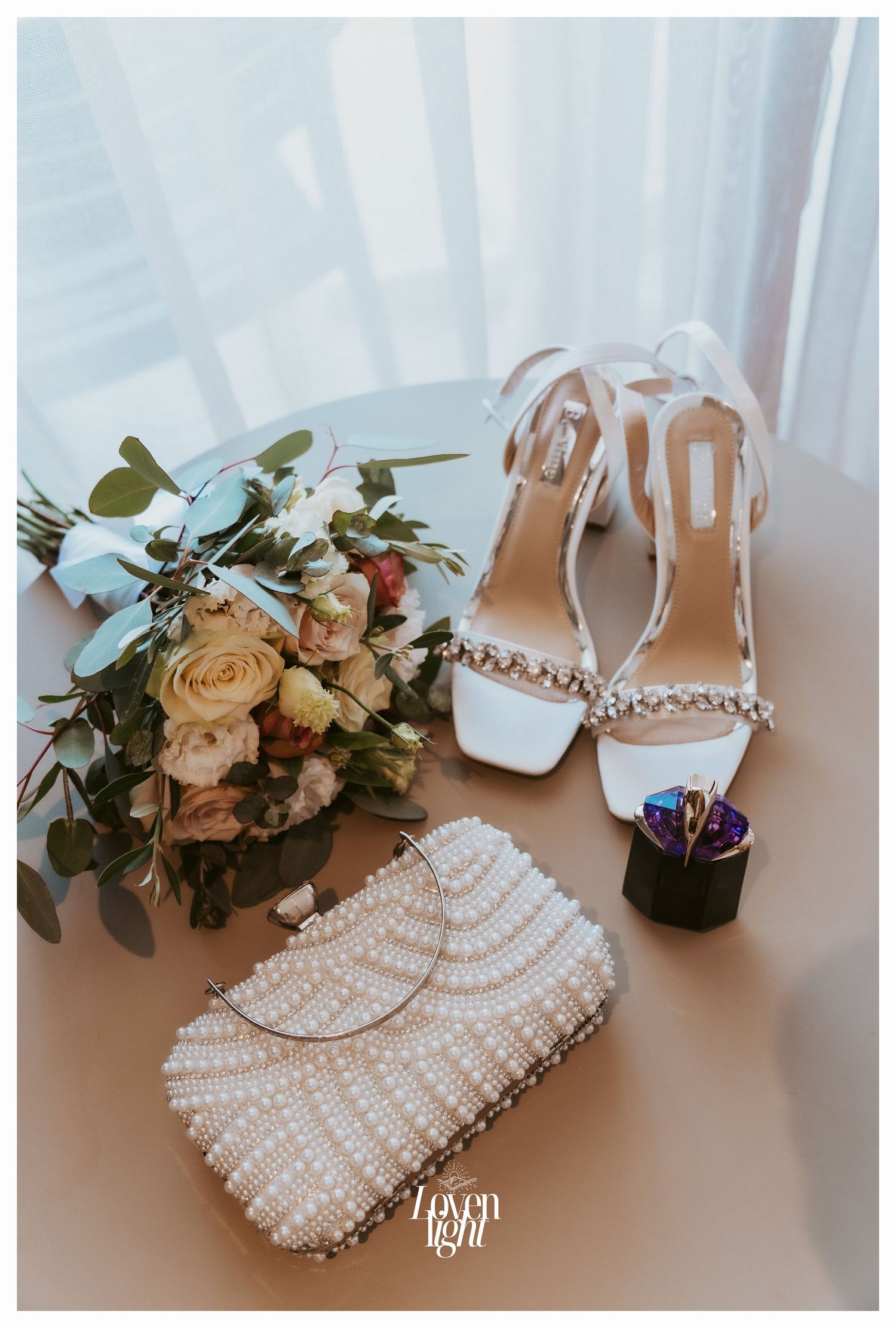 Bridal accessories including white heels, pearl clutch, and blush rose bouquet arranged on table.