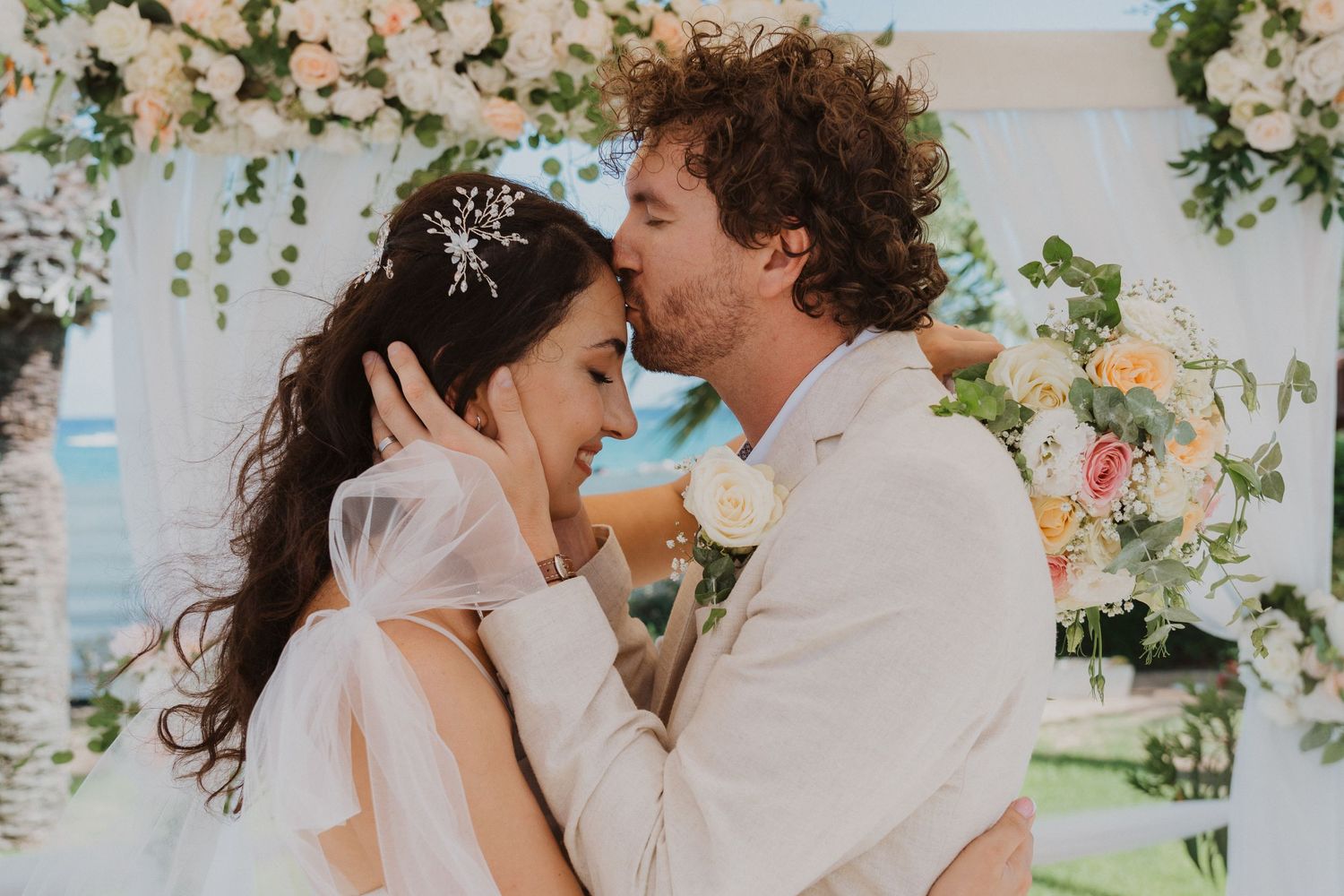 Romantic wedding moment captured under elegant floral arch at beachside venue.