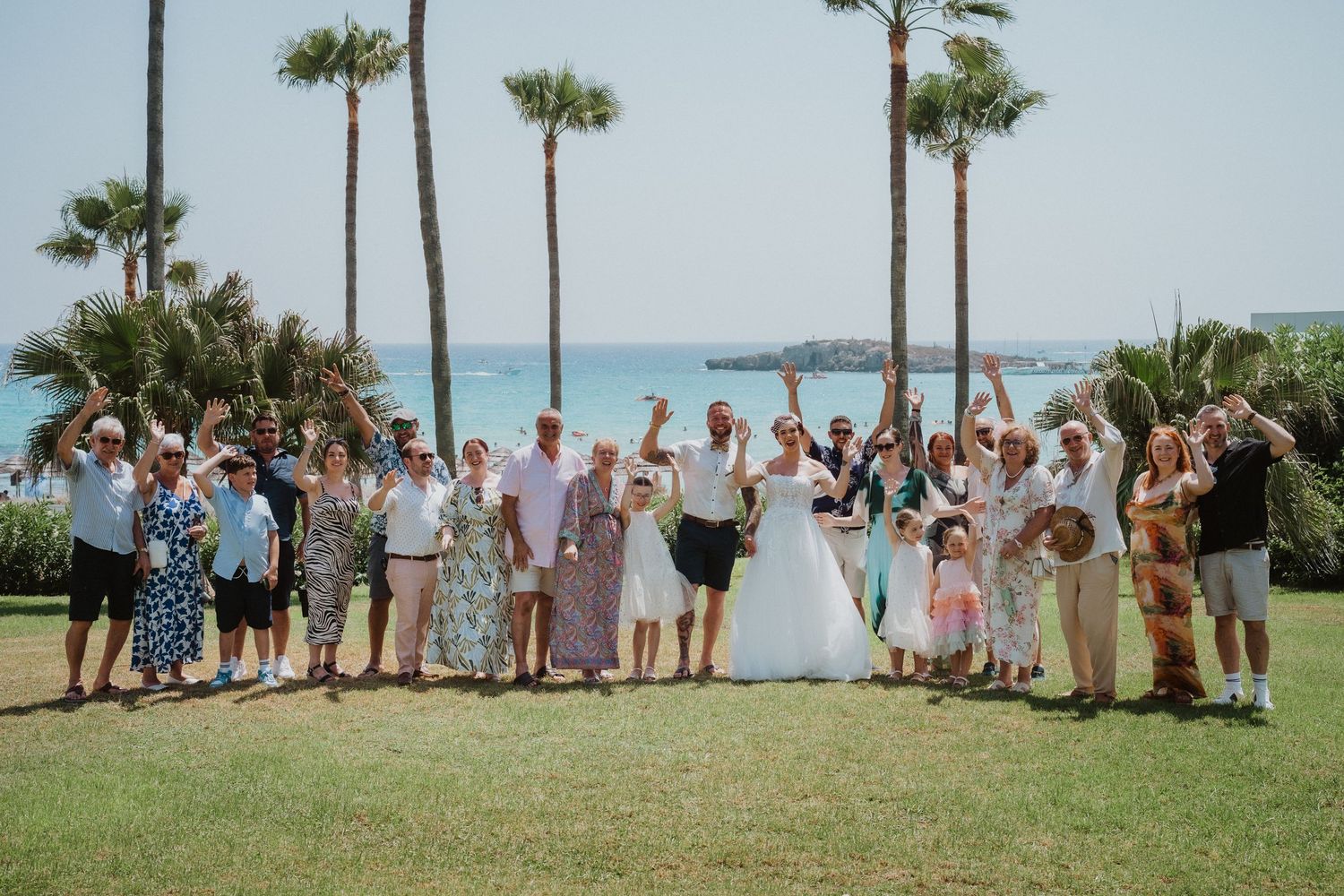 Large wedding party group gathered on a grassy lawn with tall palm trees and ocean view in the background.