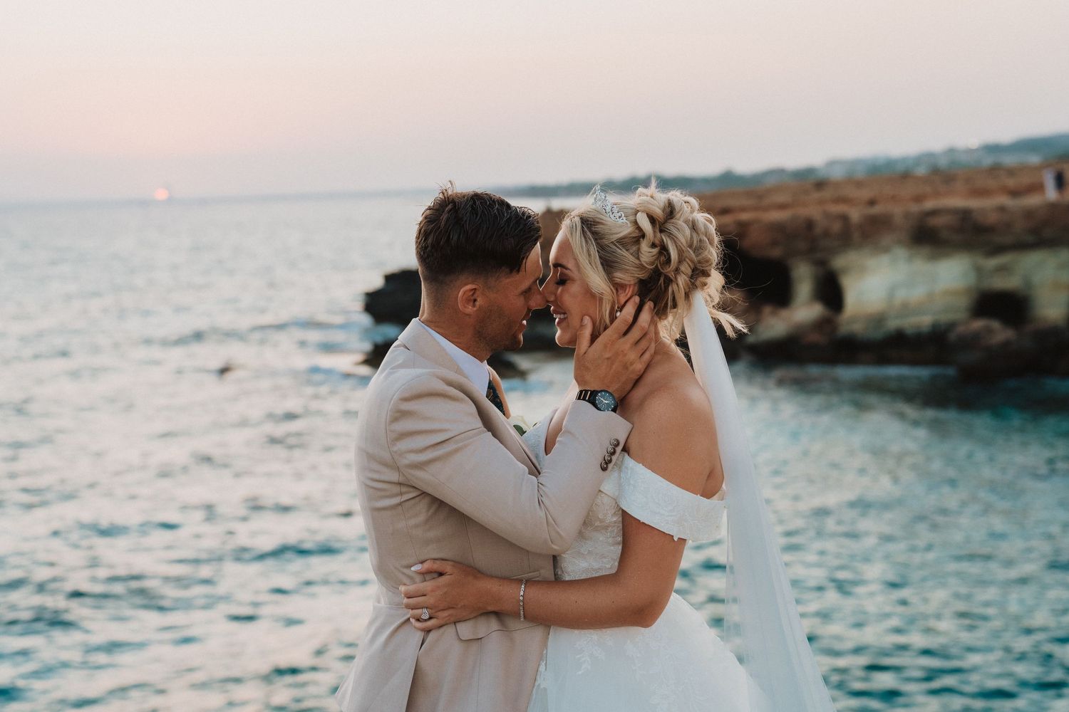 Couple in wedding attire embrace during sunset by rocky oceanside cliffs in Cyprus.