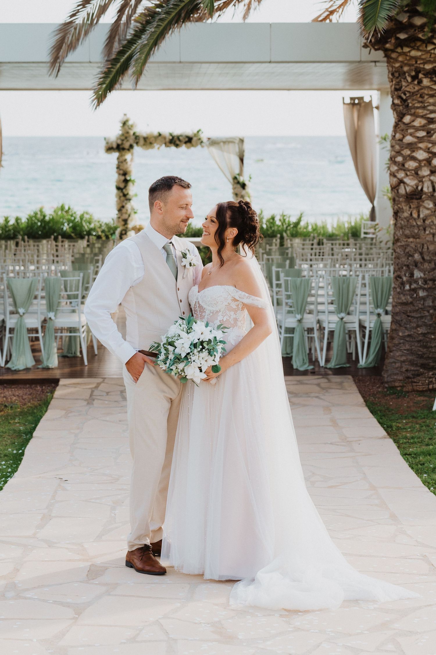 A romantic wedding couple poses by the sea under a decorated arch with palm trees and ocean views in the background.