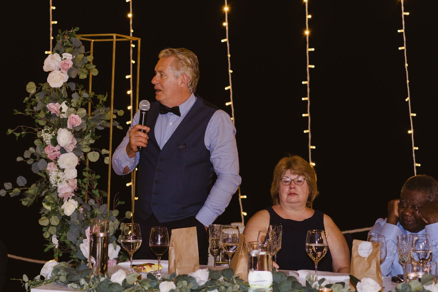 Speaker giving toast at elegant wedding reception with floral arrangements and candlelight.