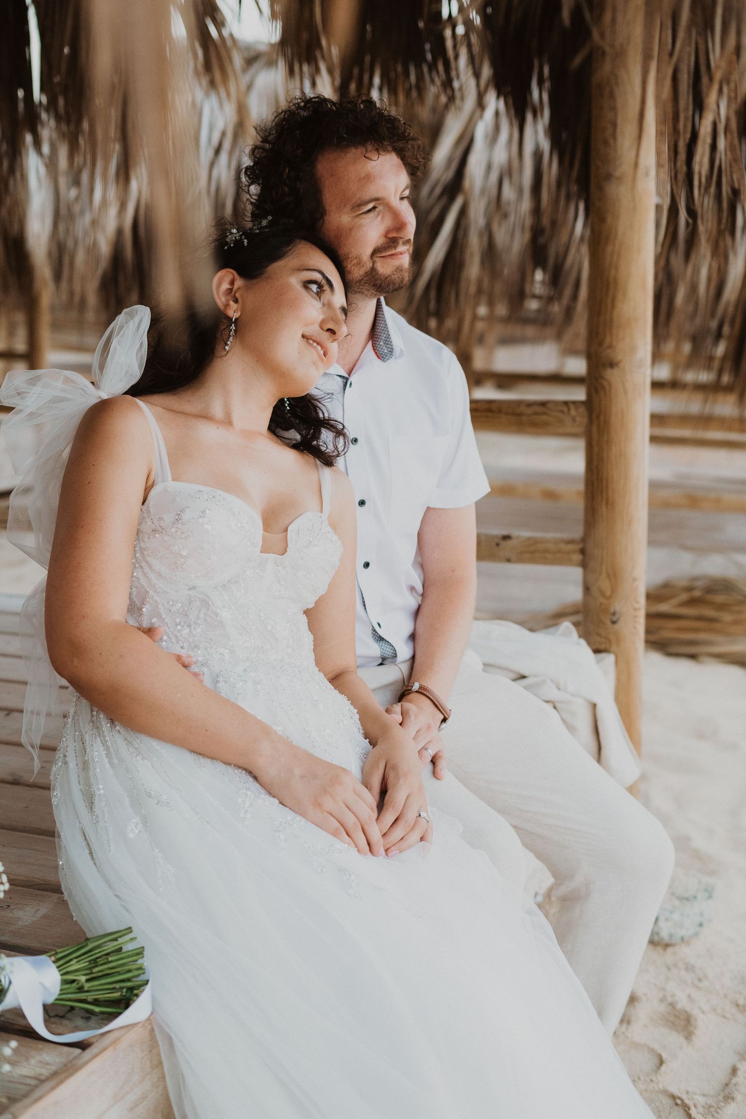A couple in white beach wedding attire shares a tender moment under a thatched palapa.
