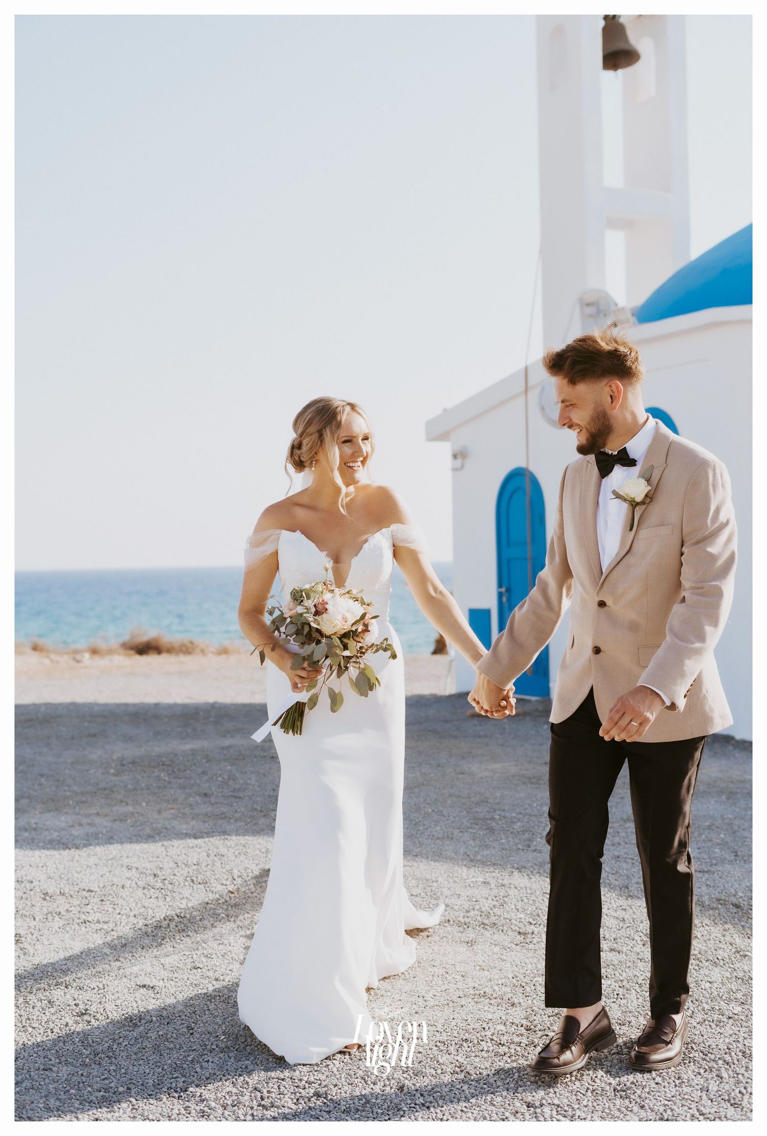 Wedding couple shares tender moment outside white Greek orthodox church by the sea.
