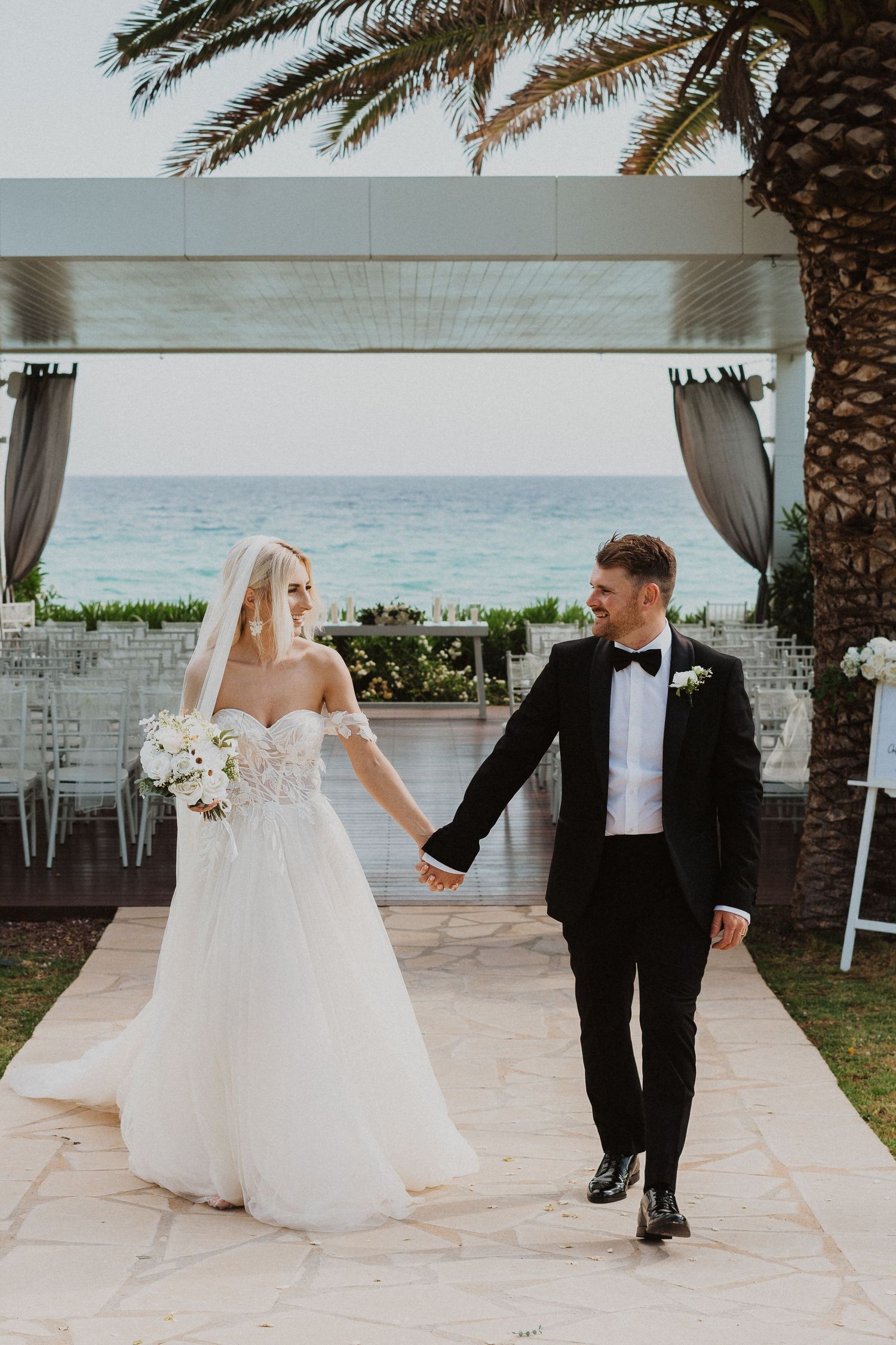 Wedding couple strolls together on a paved pathway with tropical palm trees and ocean backdrop.