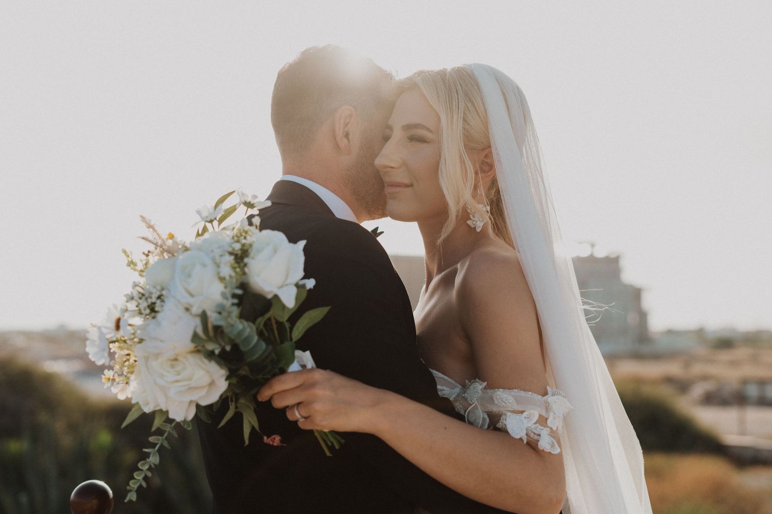 Newlyweds share an intimate moment with white bouquet against soft sunlit backdrop.