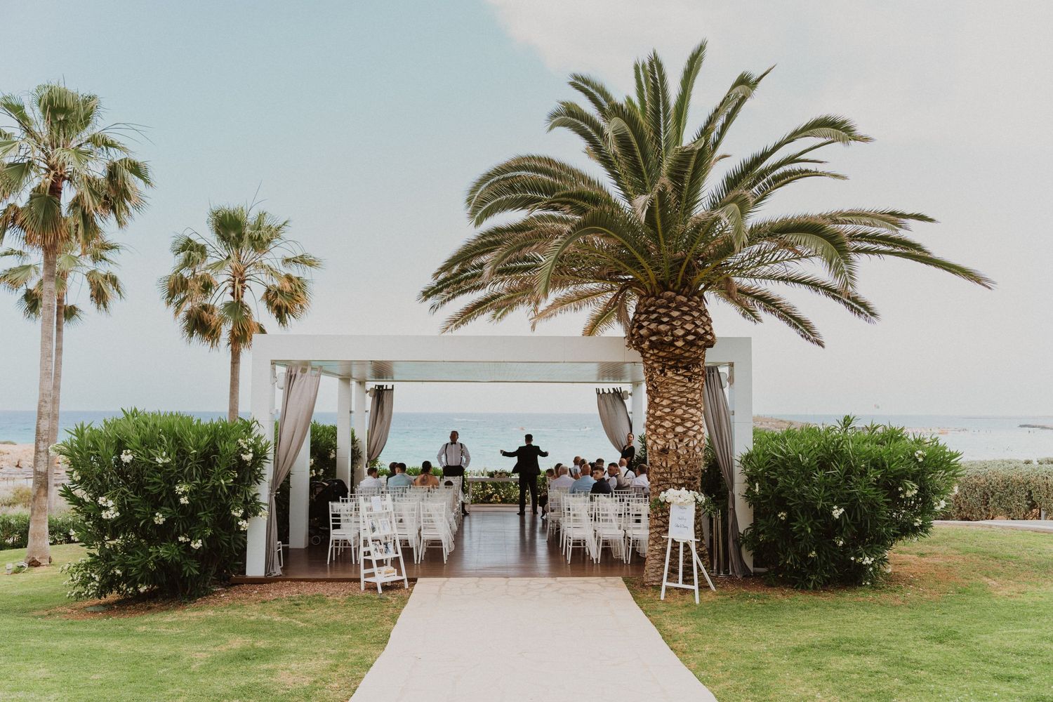 Outdoor wedding ceremony setup with white decor and palm trees at a tropical beachside venue.