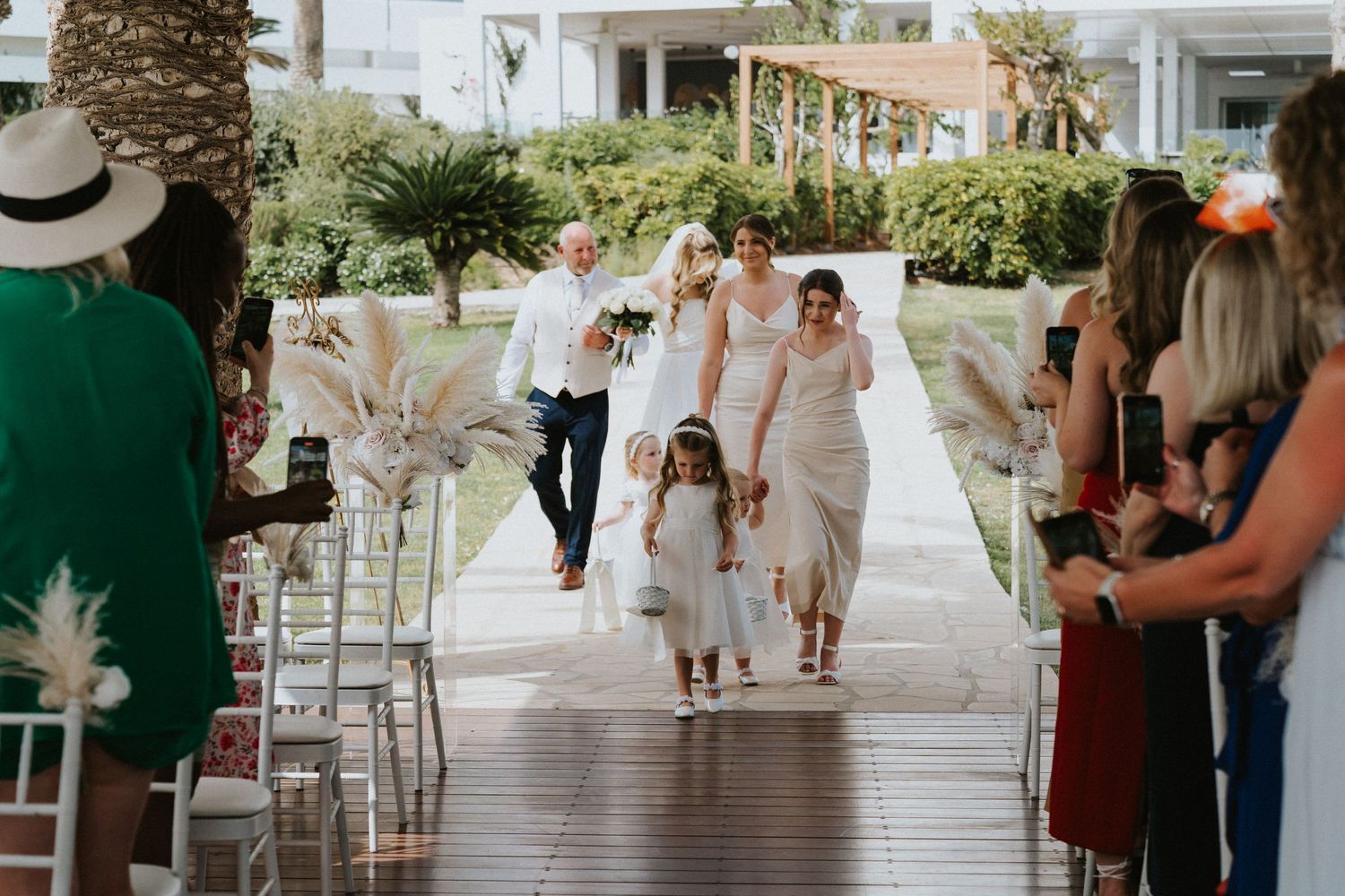 Wedding party walks down covered outdoor walkway lined with white chairs and tropical landscaping.