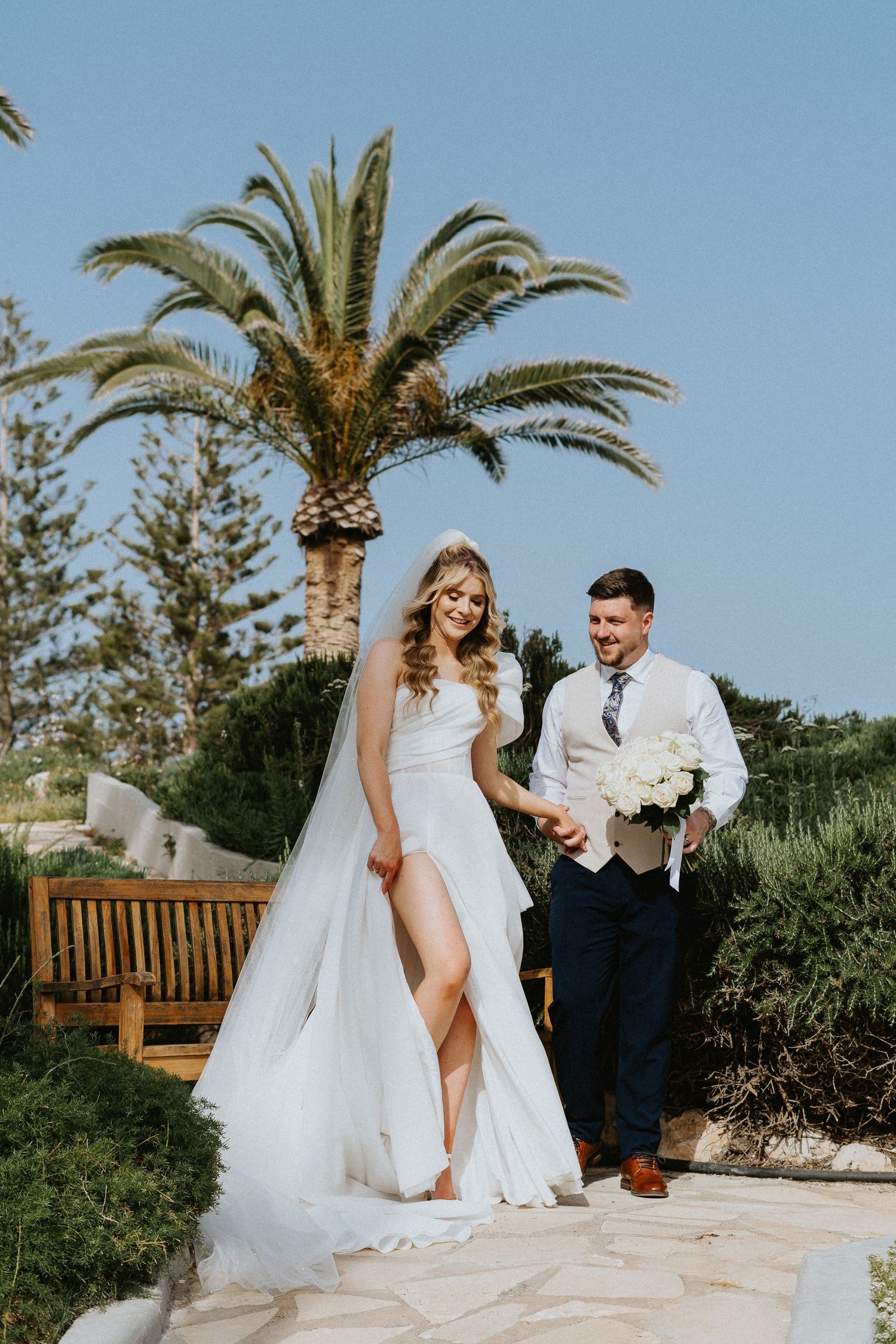 A couple in wedding attire pose together on a stone pathway with a palm tree and blue sky in the background.