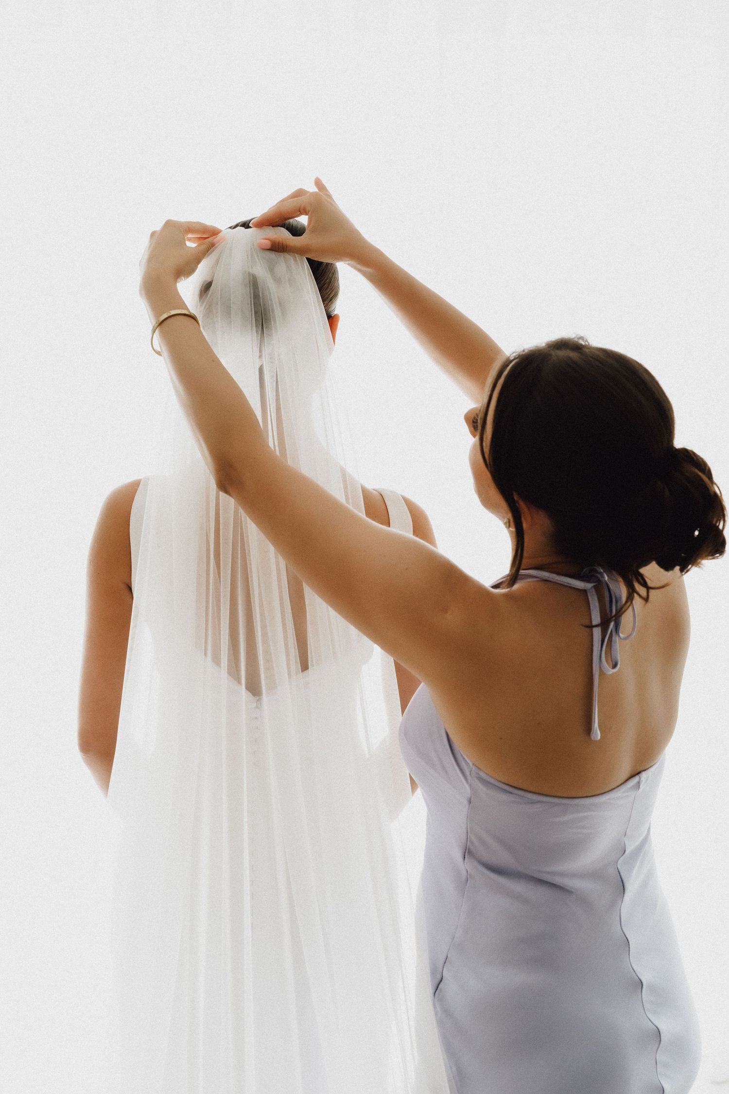 Silhouetted figure helping to adjust a flowing white wedding veil in natural light.