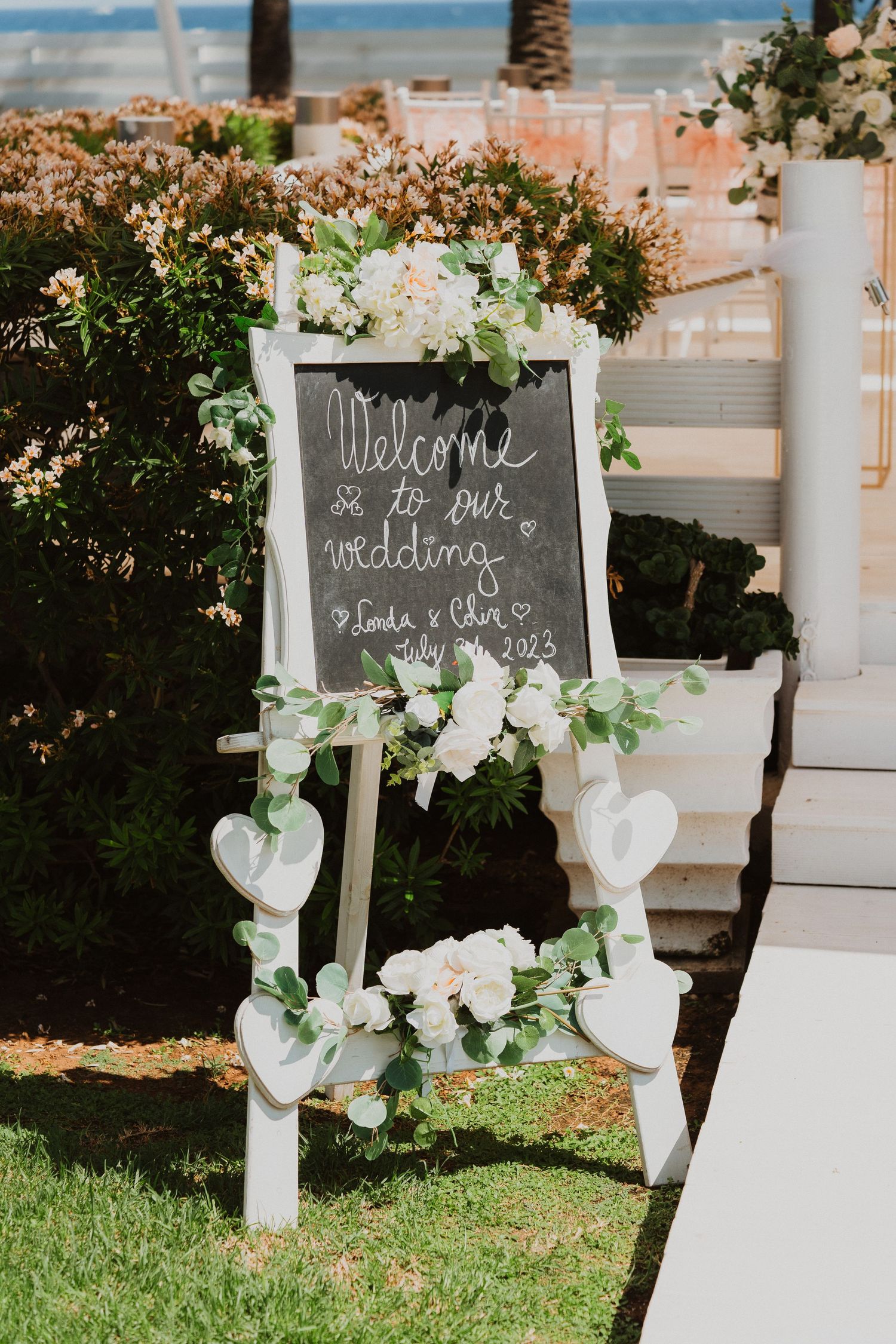 A rustic chalkboard welcome sign decorated with white flowers and heart ornaments stands on grass at an outdoor wedding venue.