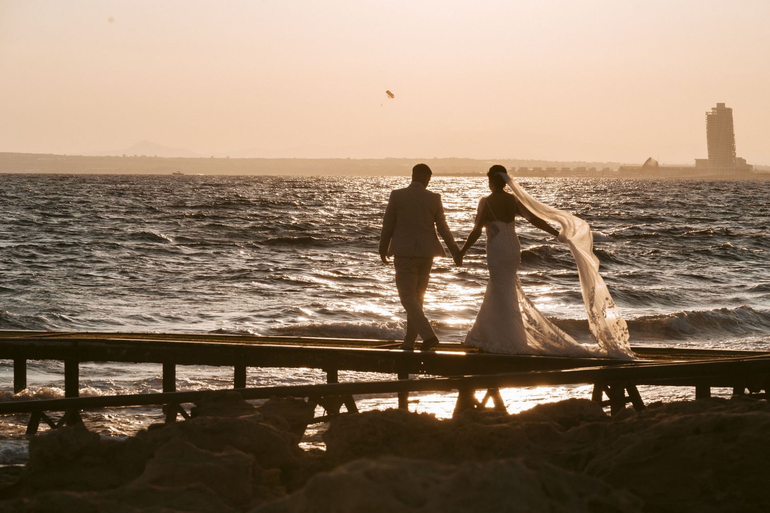 Silhouetted couple stands on pier during sunset with ocean waves crashing.