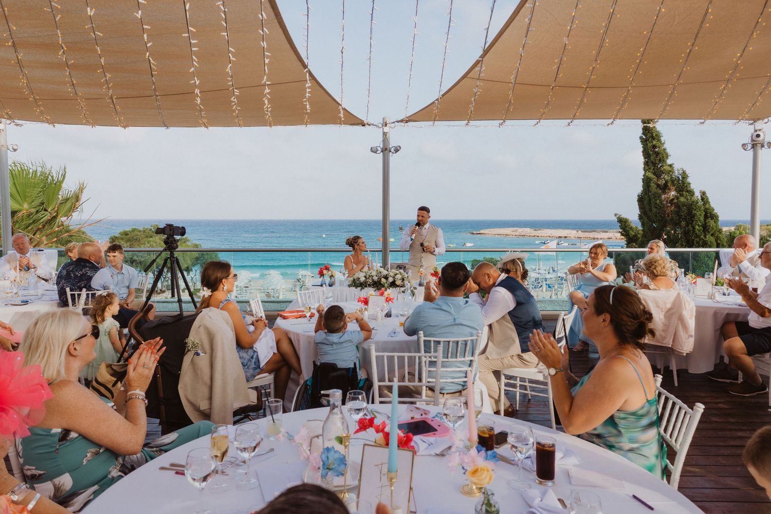 Guests enjoying a beachside wedding reception under a white tent canopy with ocean views in the background.