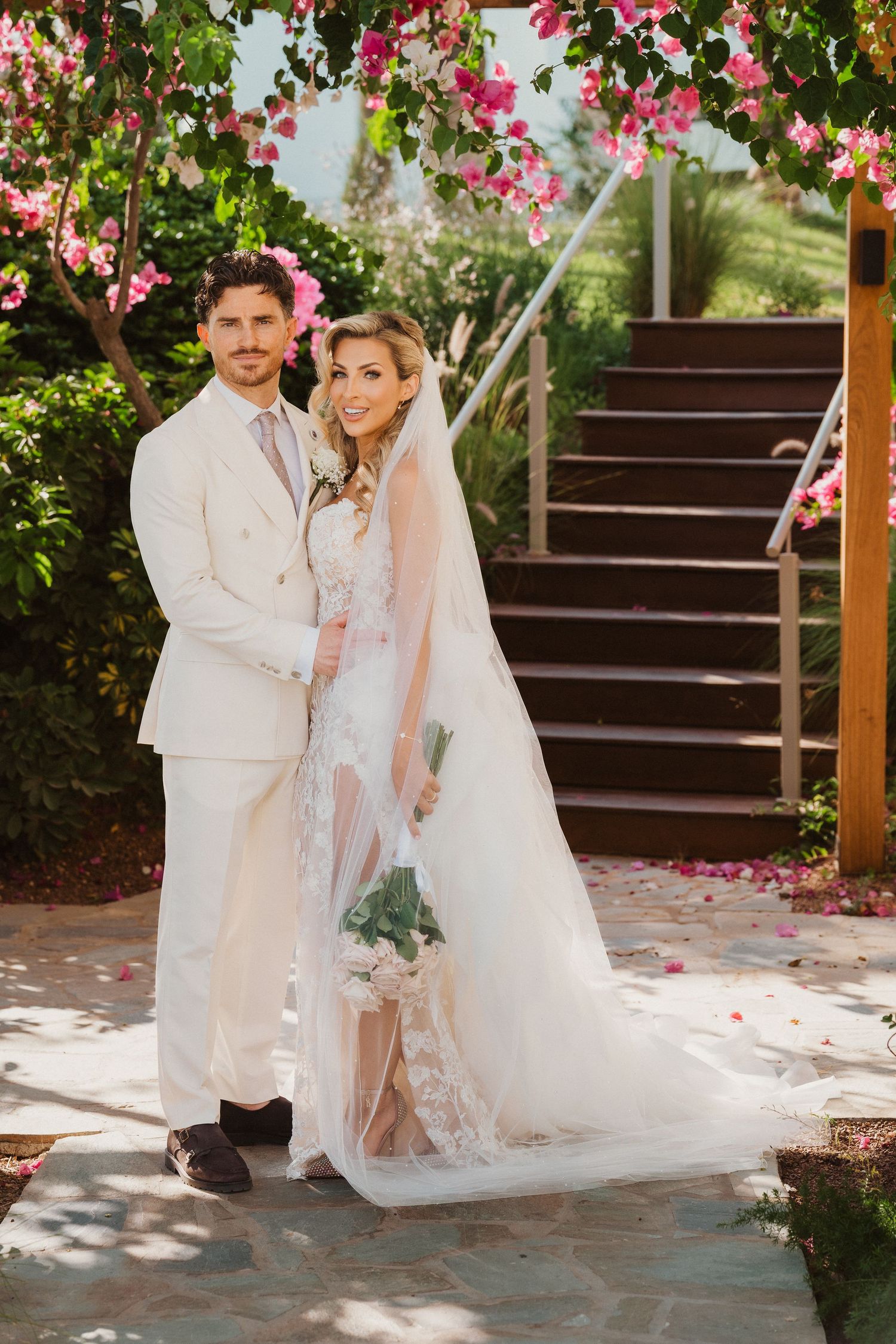 A wedding couple poses under pink floral archway on garden steps wearing elegant white and cream attire.