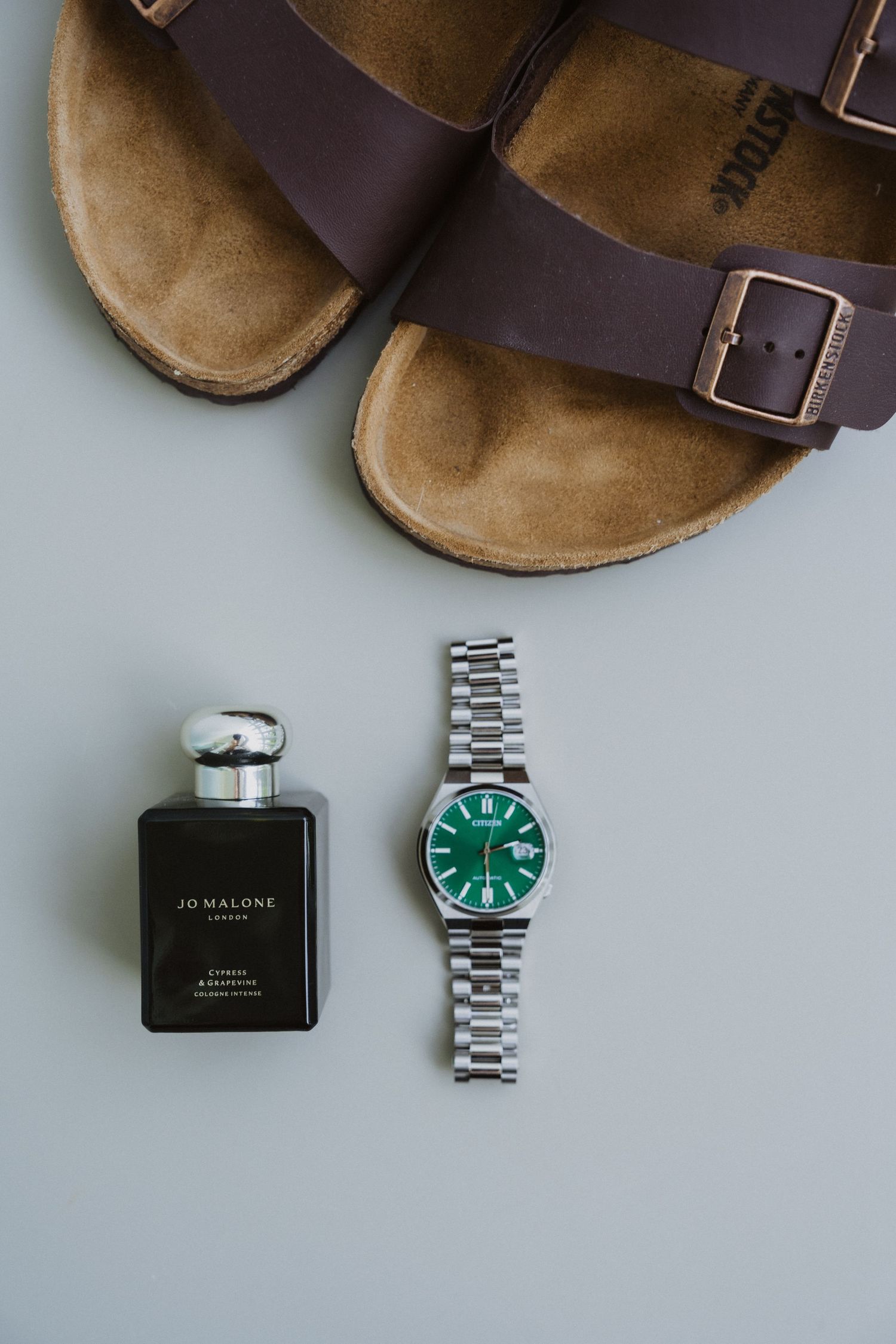 Brown Birkenstock sandals with Jo Malone perfume and green-faced watch on gray surface.