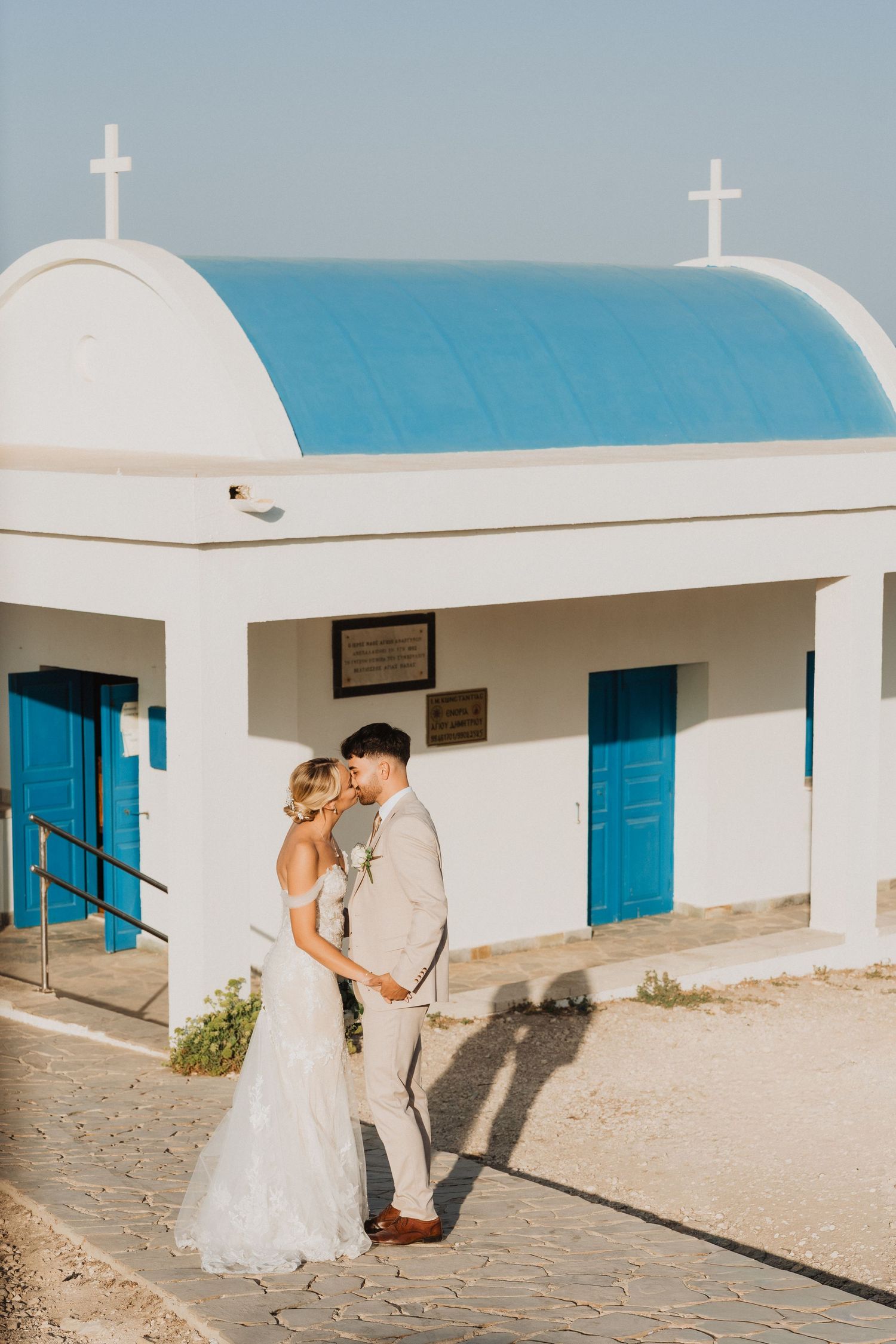 A wedding couple poses in front of a traditional white Greek chapel with blue doors by the sea.