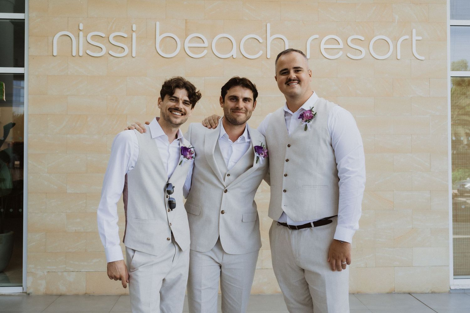 Three men in light colored suits smile together at the Nissi Beach Resort entrance.