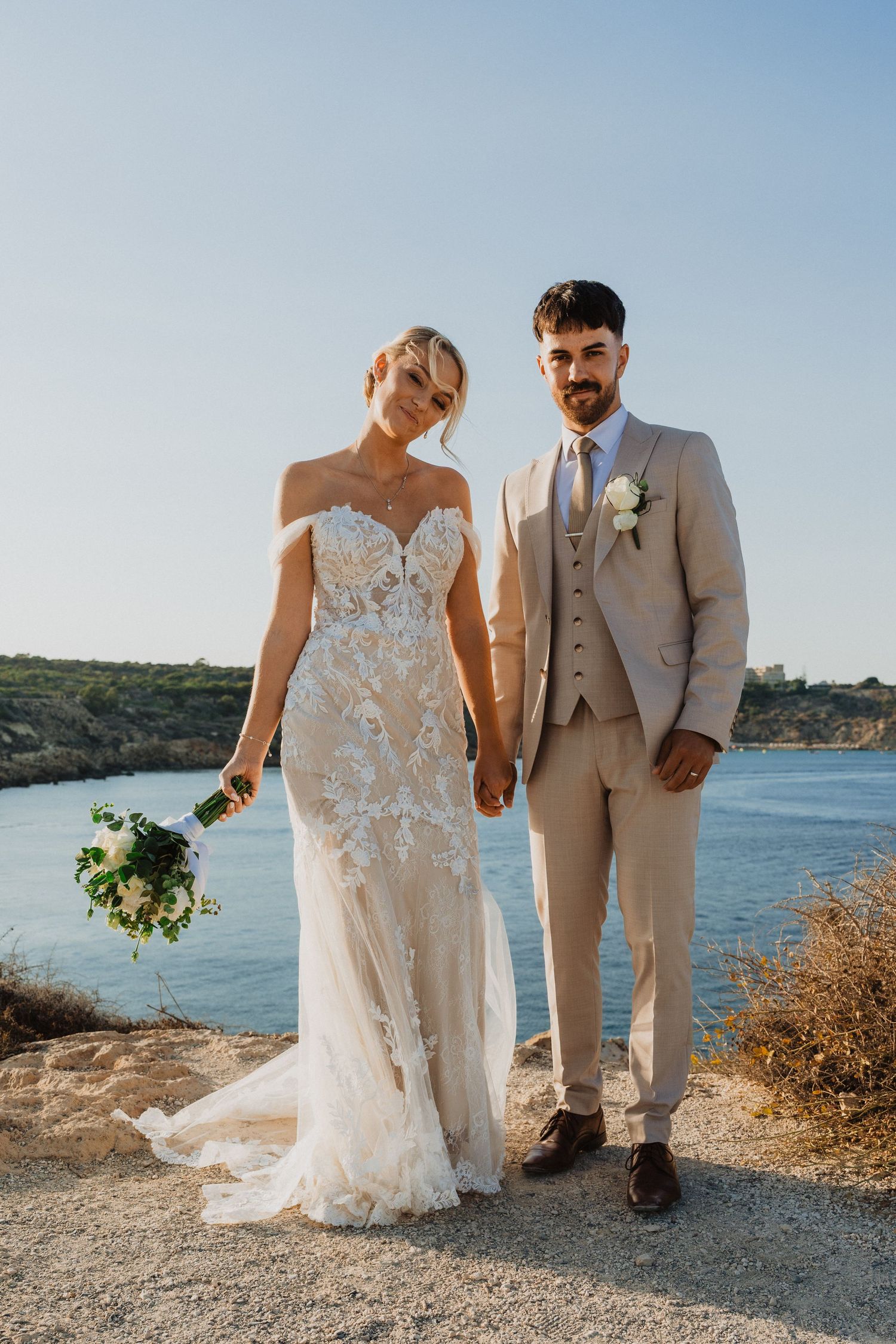 Elegant wedding couple in lace gown and beige suit posing against coastal landscape.