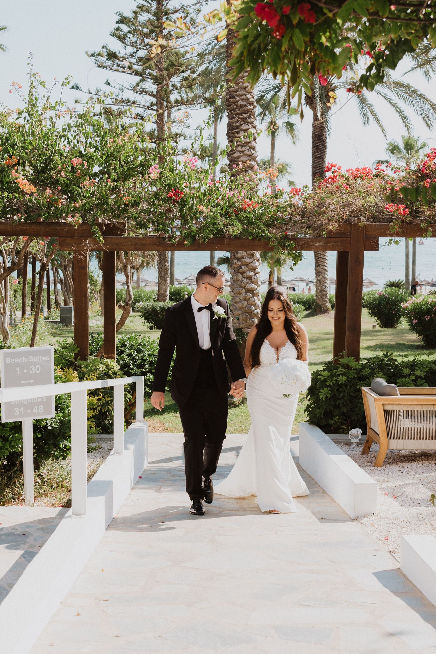 A couple walks along a white pergola walkway decorated with tropical flowers and palm trees.