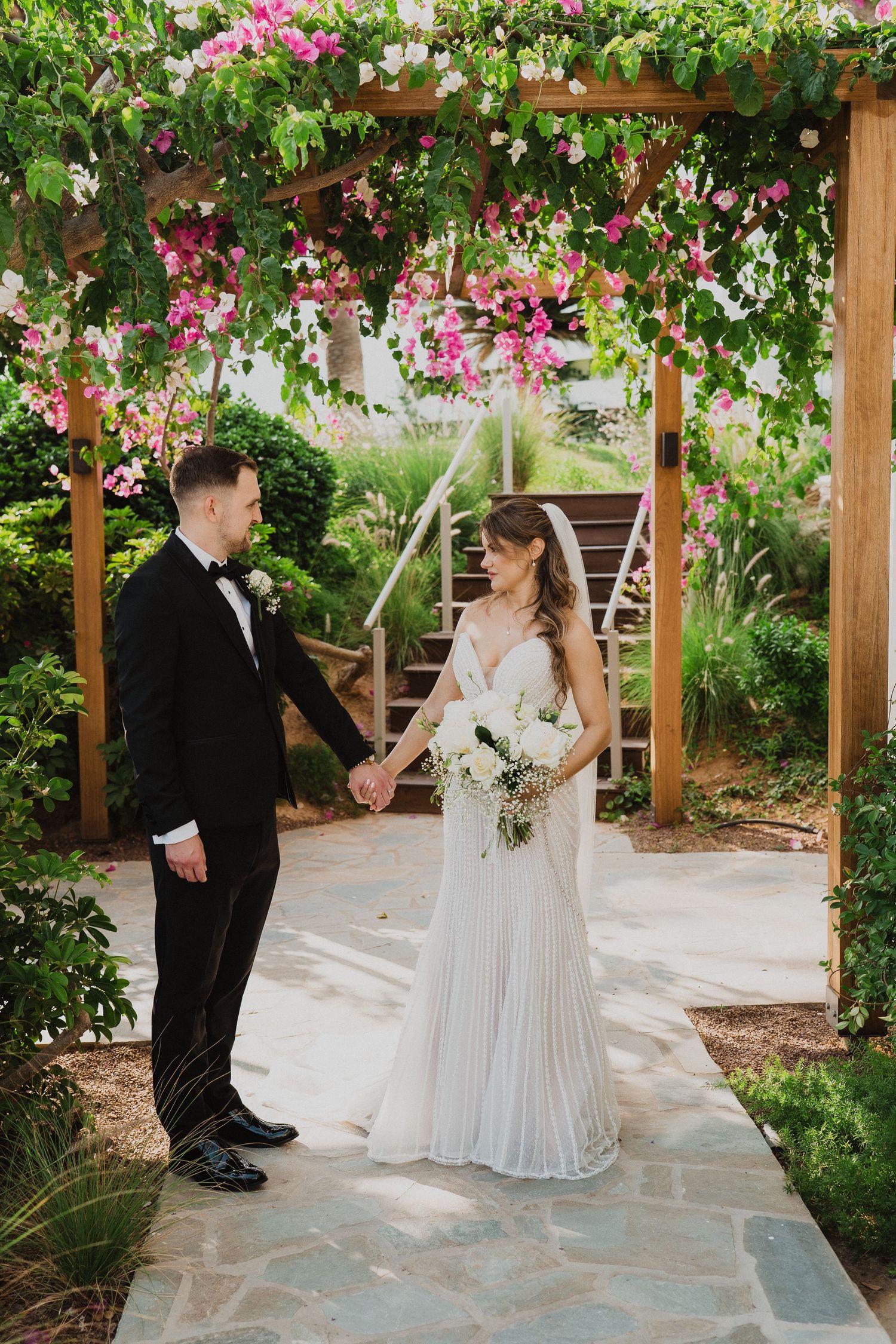 Bride and groom hold hands under a floral pergola with pink and white bougainvillea at an outdoor wedding.