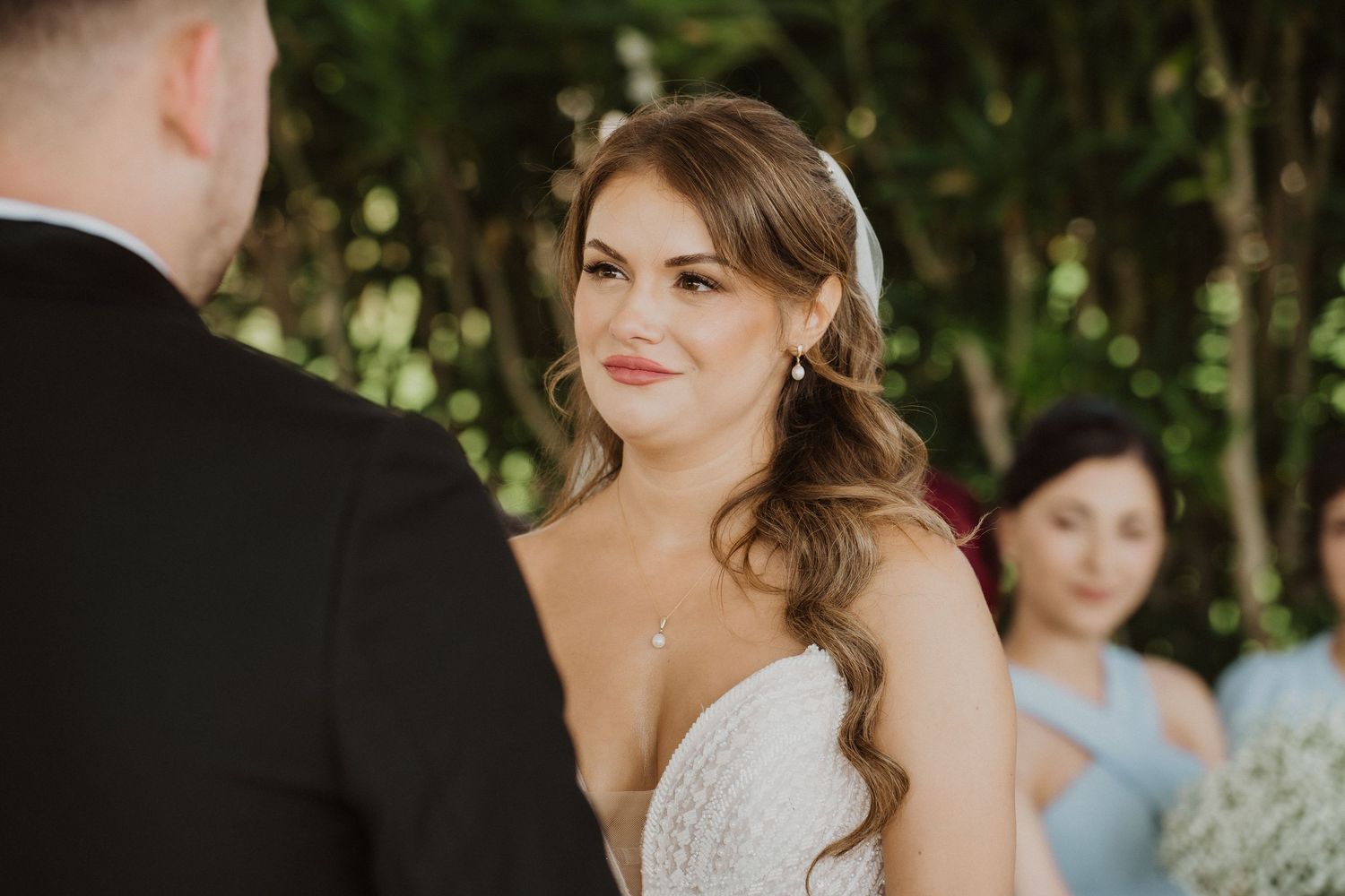 Bride smiling warmly at groom during outdoor wedding ceremony with bridesmaids behind.