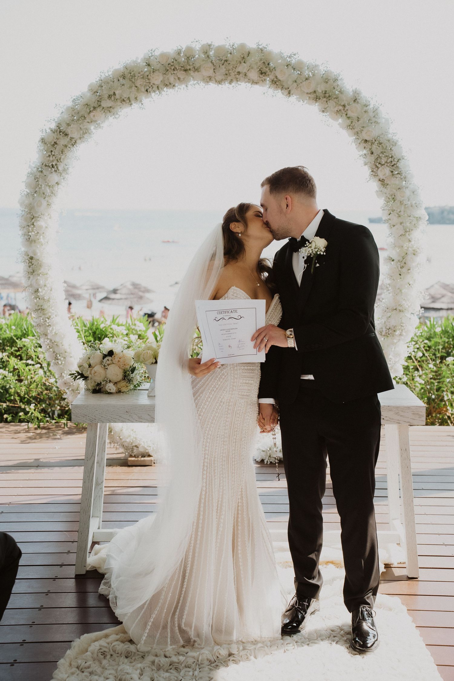 Bride and groom share first kiss under floral circle arch at outdoor beach wedding ceremony holding marriage certificate.