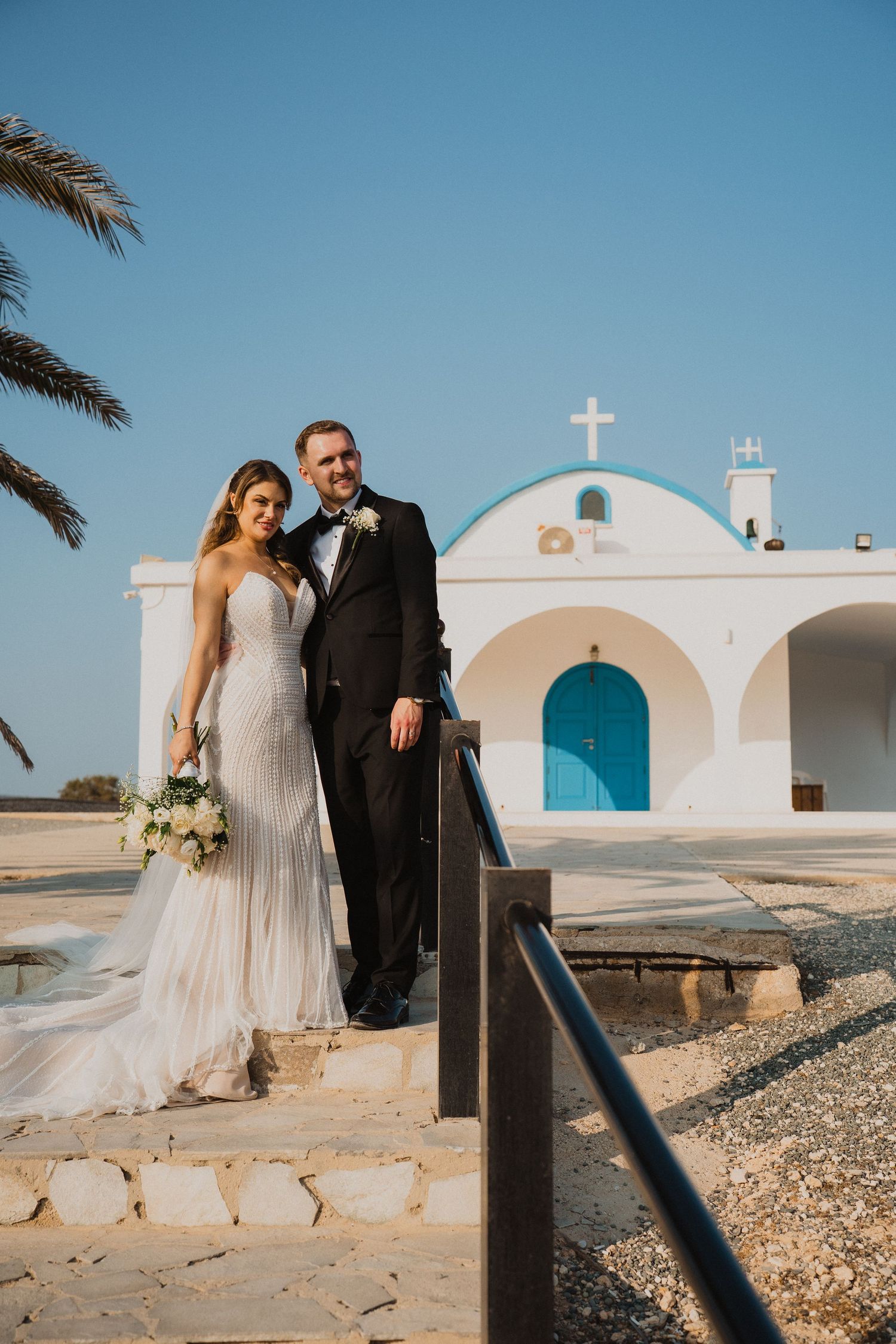 Bride and groom posing before a white Greek chapel with blue dome and door in sunny Cyprus.