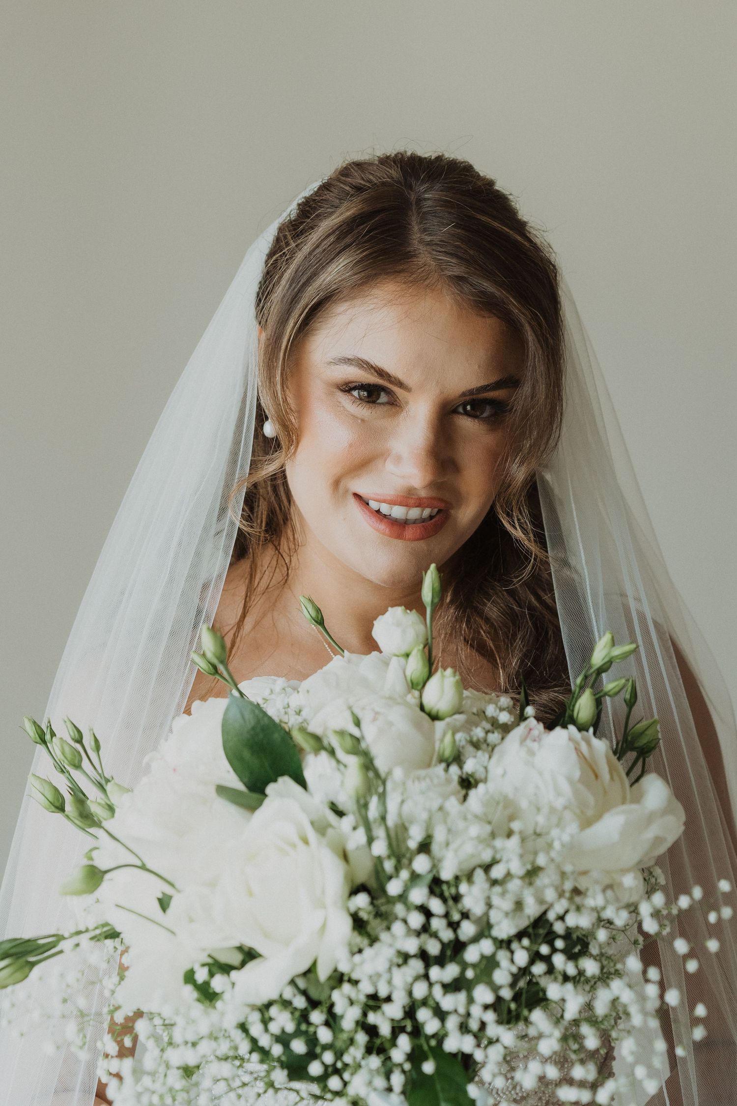 Beautiful bride smiling with white floral bouquet and veil in soft natural light wedding portrait.