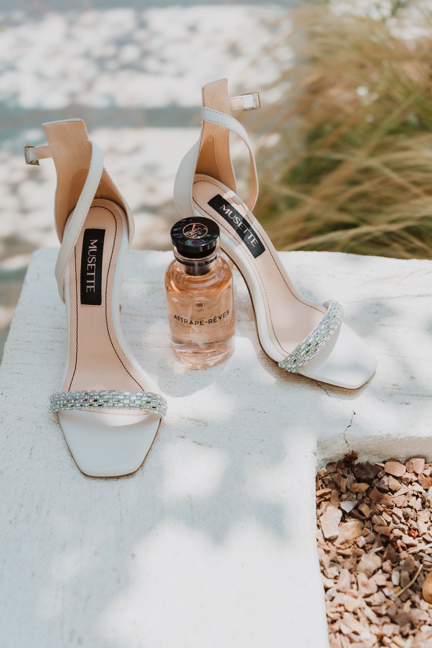 Elegant white Mesette bridal heels with crystal straps beside a luxury perfume bottle on a white surface outdoors.