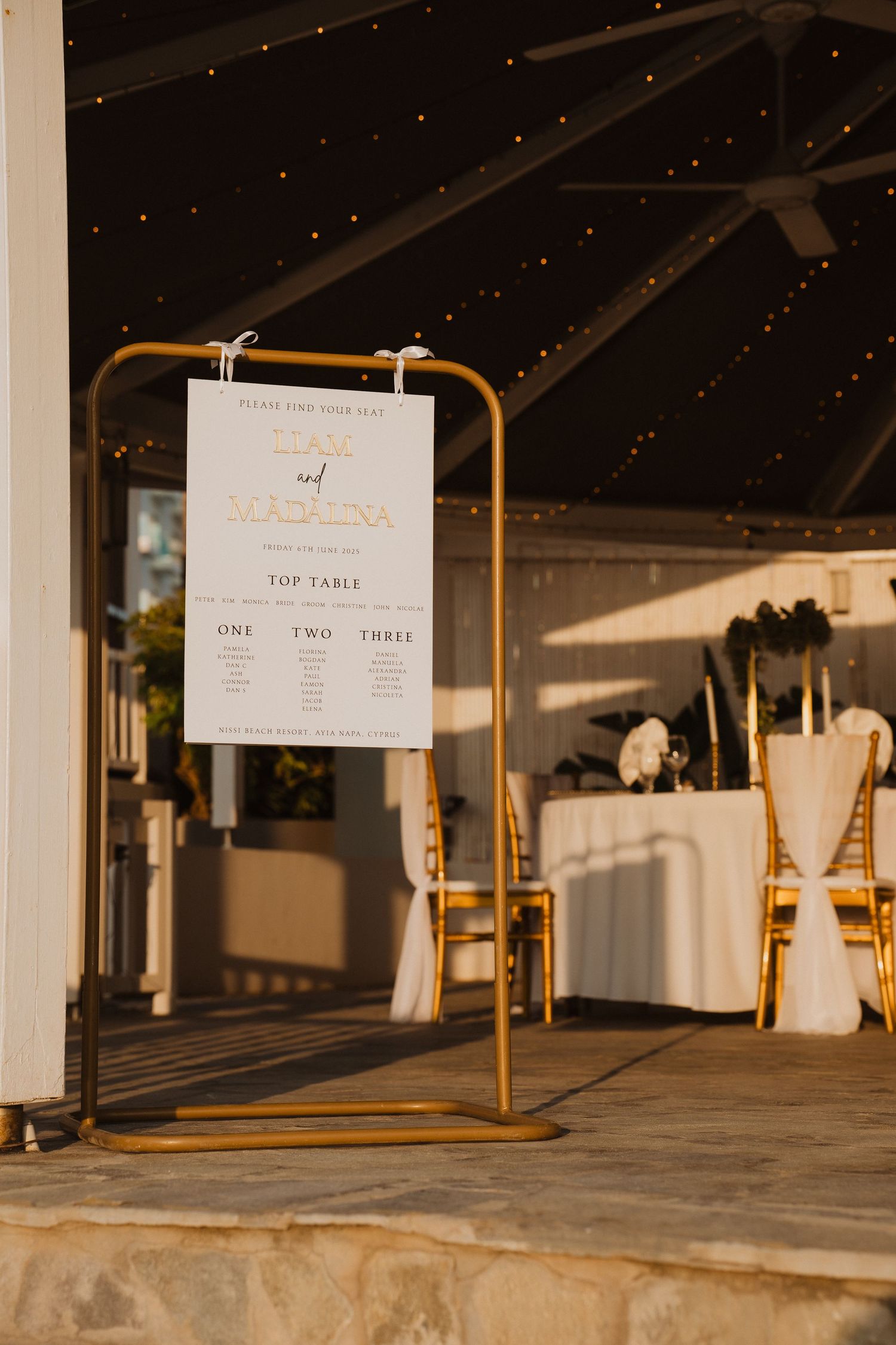 Gold standing seating chart display at elegant outdoor wedding reception venue with twinkling string lights.