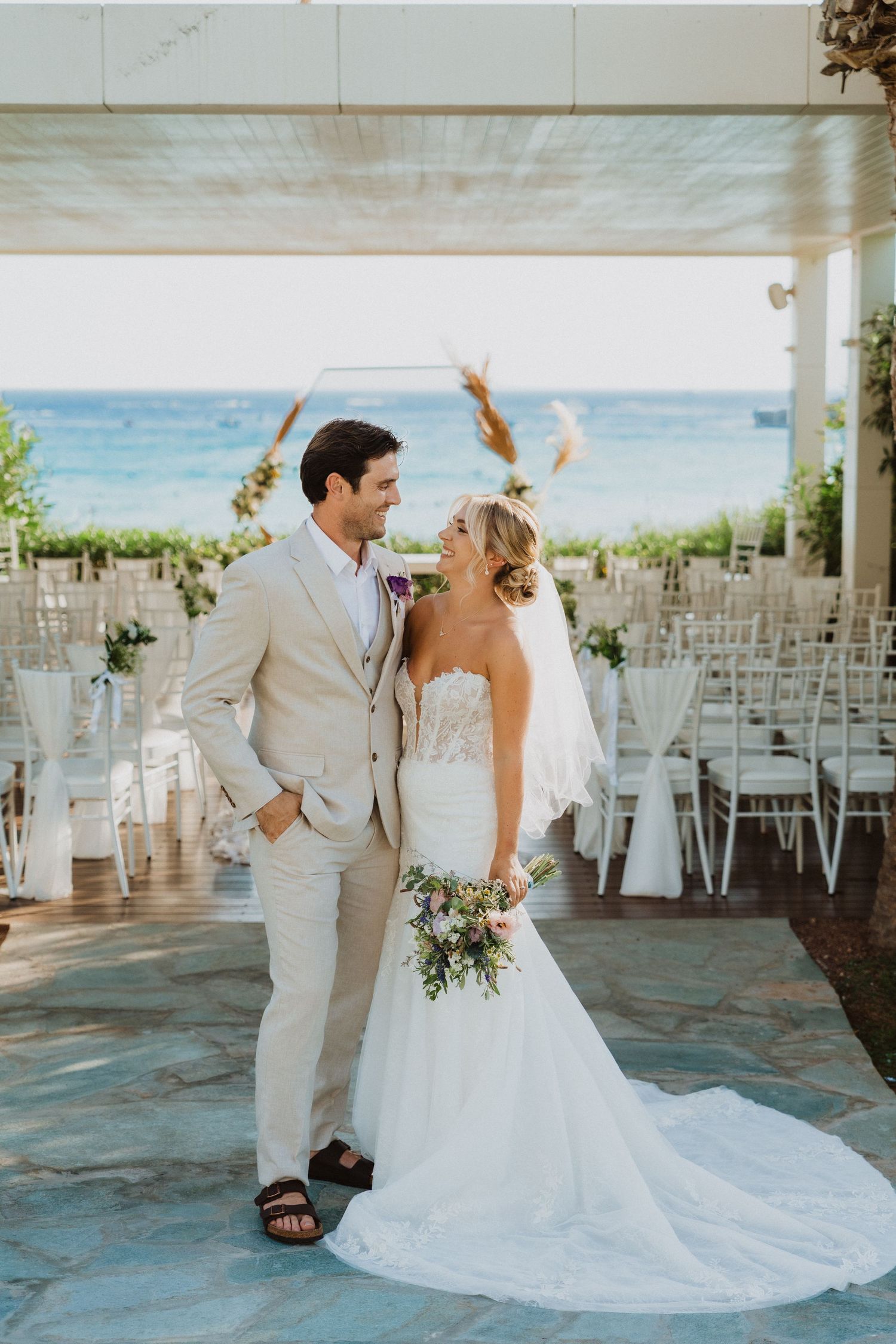 A romantic wedding couple shares an intimate moment on a beachfront patio with white chairs and ocean views.