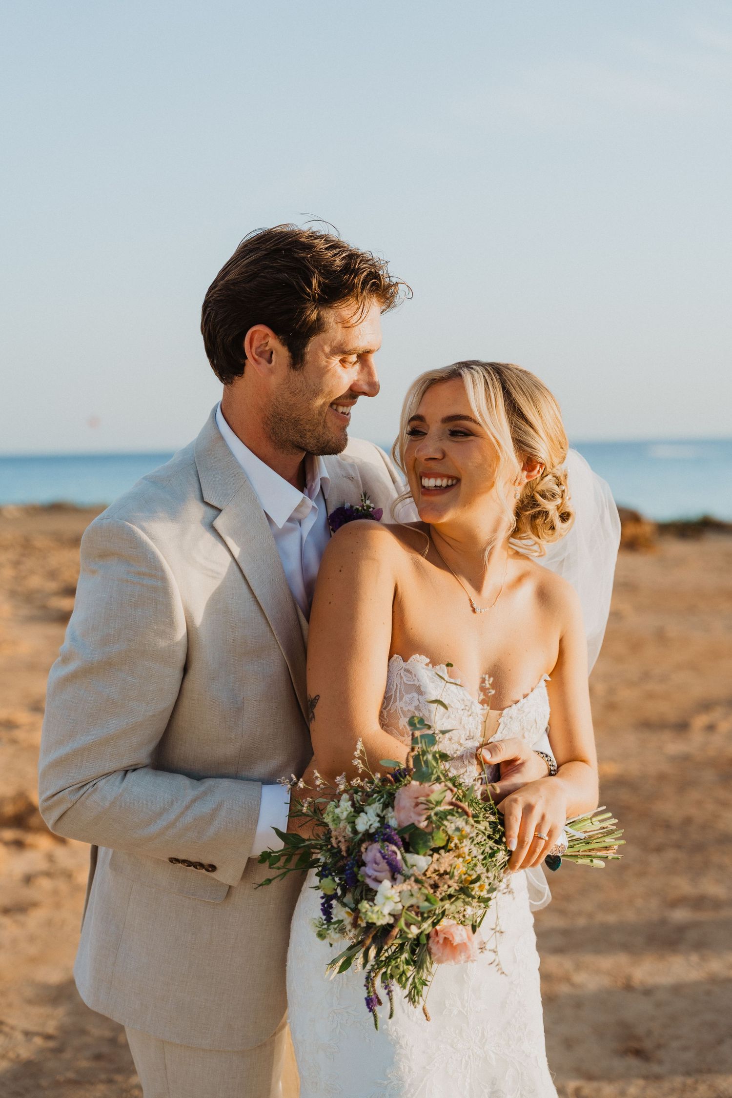 Joyful wedding couple laughs and embraces while holding a rustic bouquet by the seaside.
