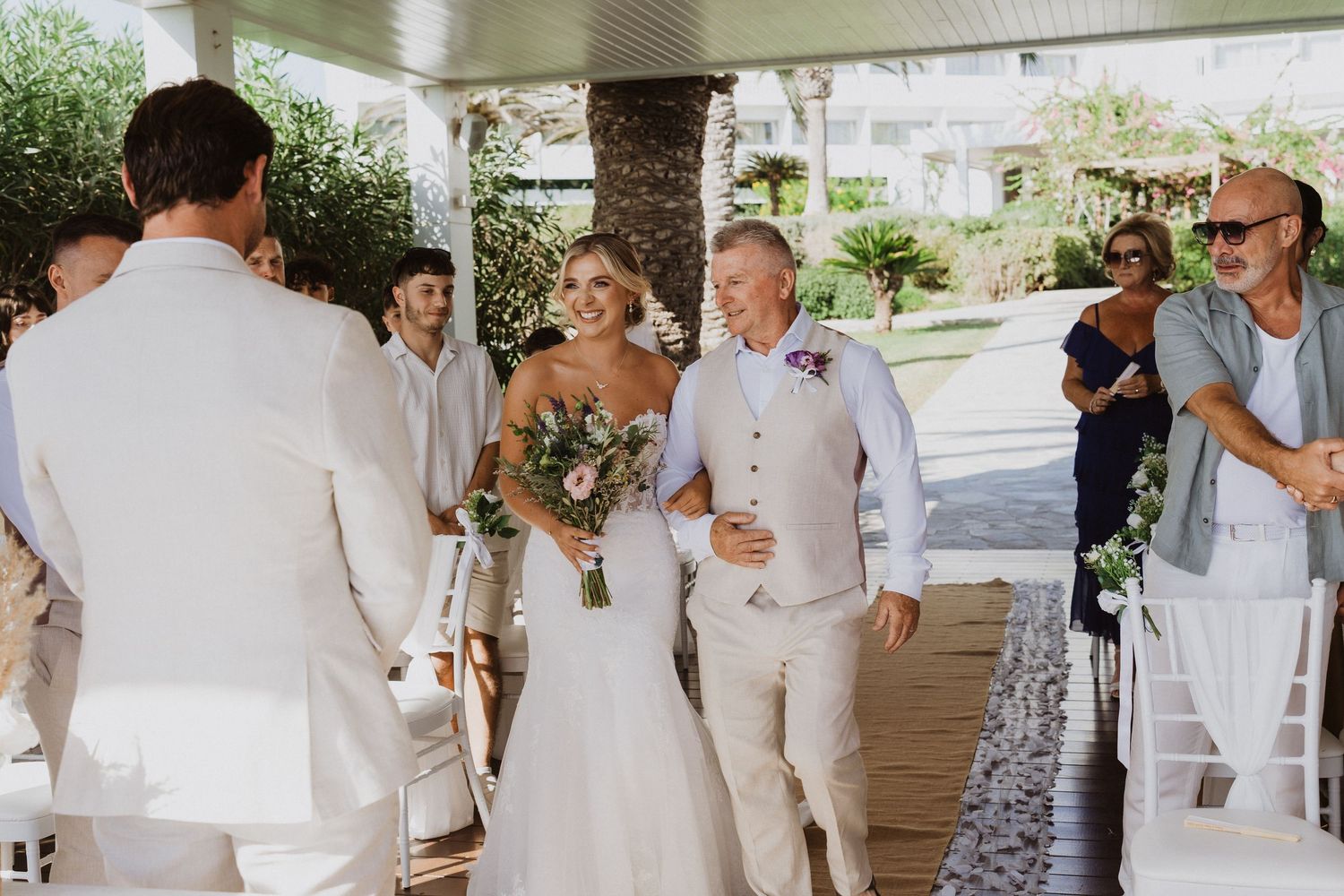 Wedding ceremony processional under a covered outdoor venue with white chairs and floral decorations.