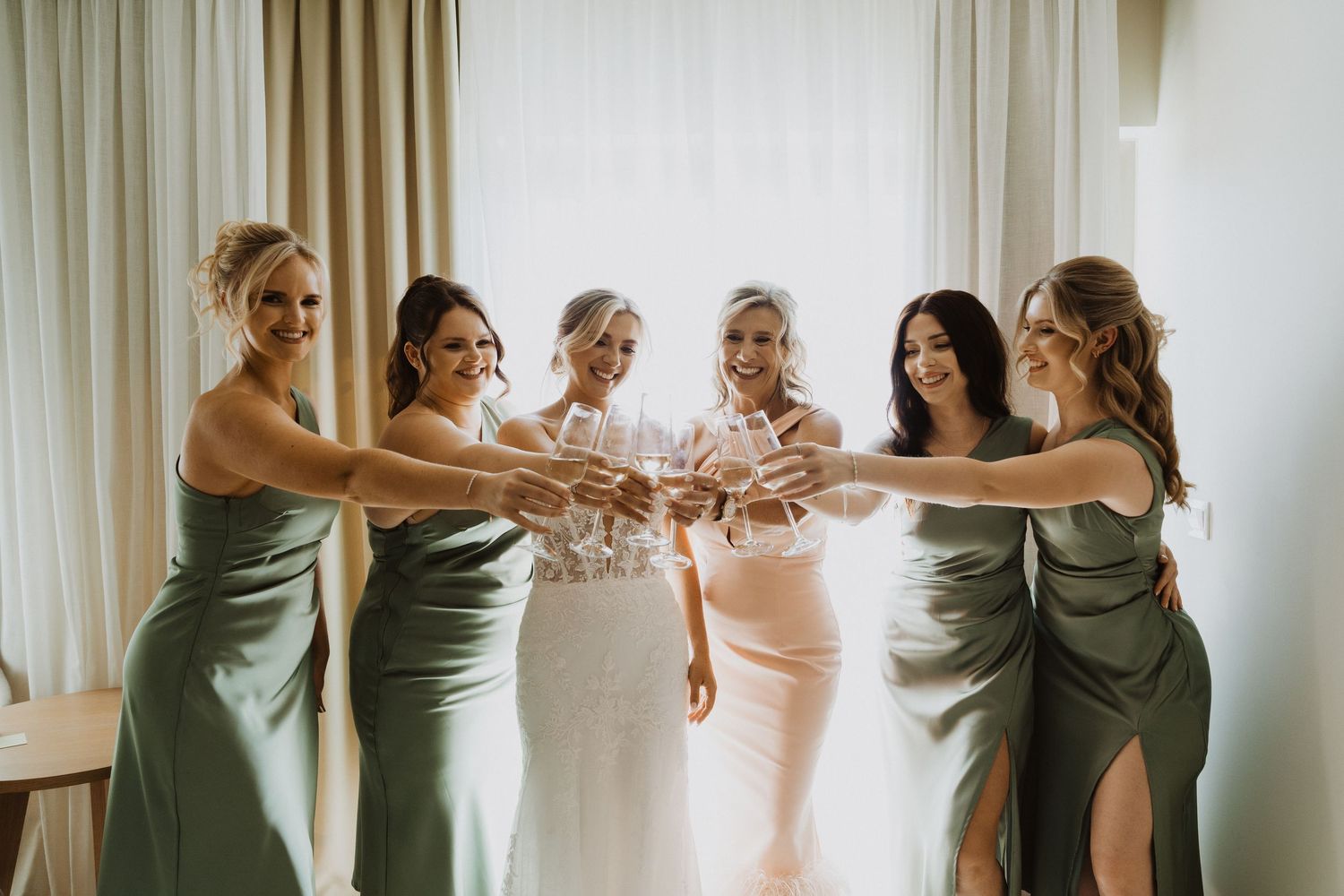 Bridesmaids in sage green and blush pink dresses gather together for a pre-wedding moment.