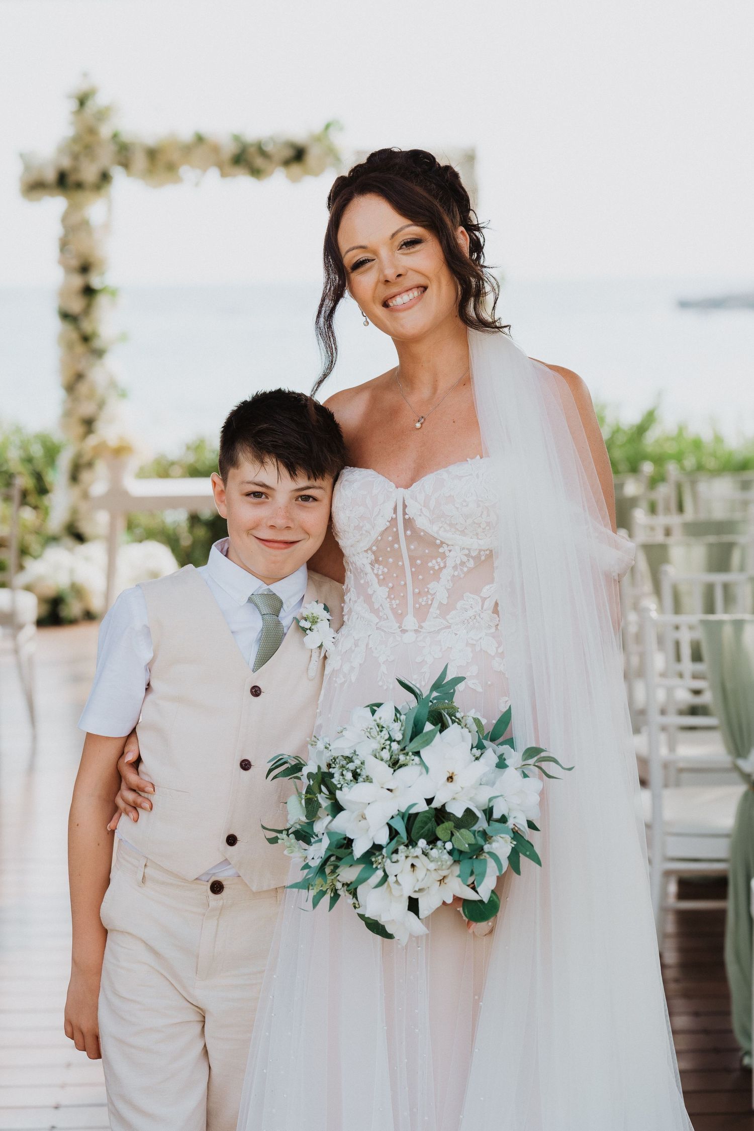 A bride in a white lace gown stands with a young boy in a beige vest at a beachside wedding ceremony with floral decor.