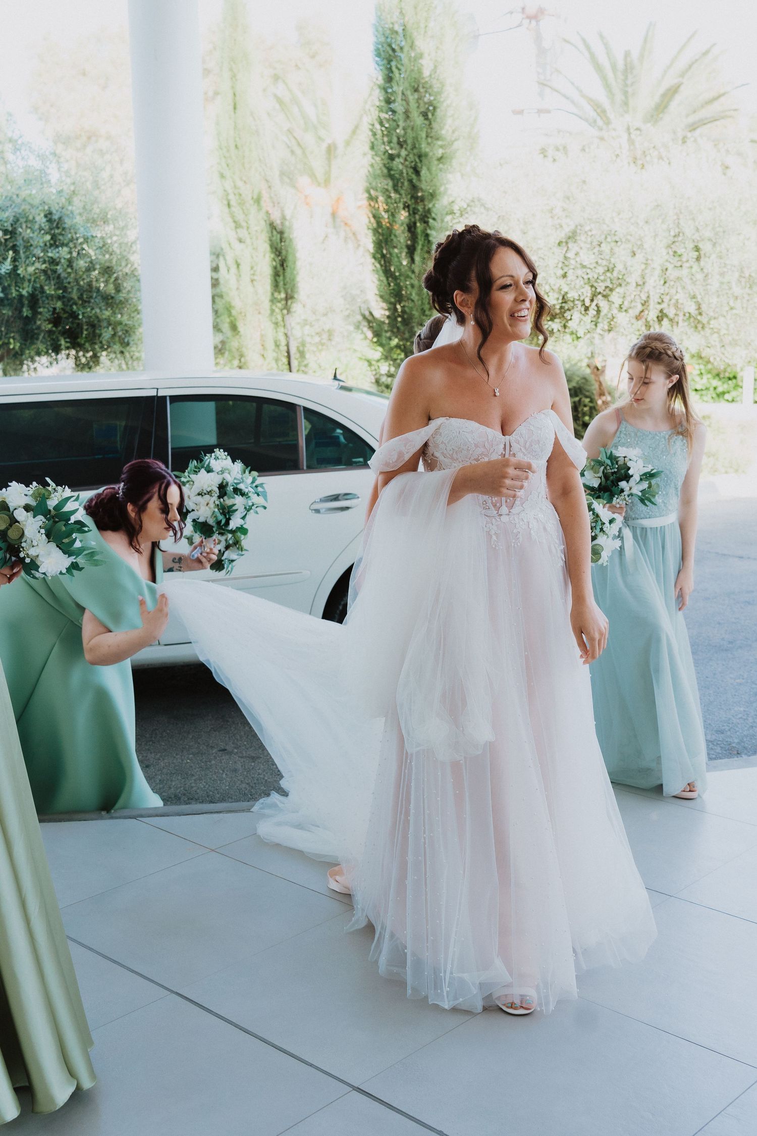 A bride in a flowing white tulle gown stands while bridesmaids in mint green dresses assist with her dress preparations.