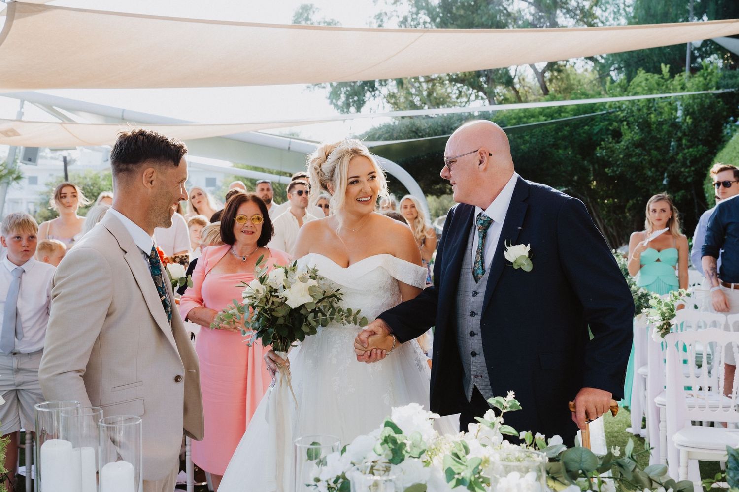 An outdoor wedding ceremony under canopy sails decorated with white flowers and floral arrangements.