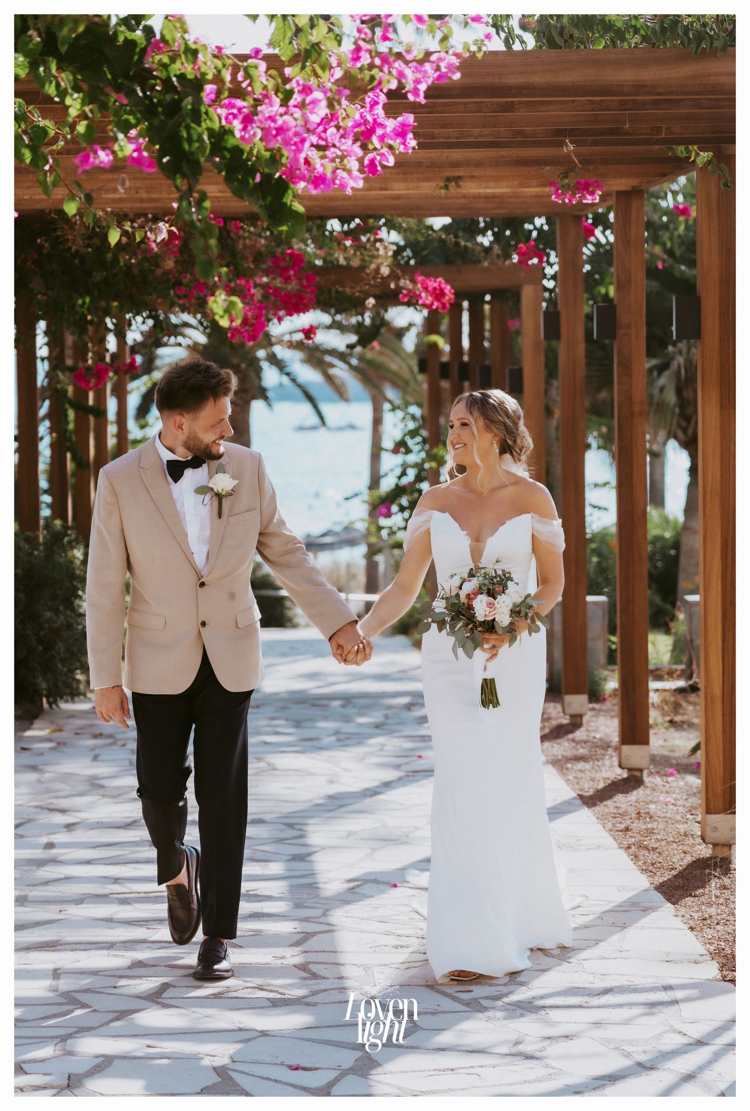 Wedding couple walking hand in hand under a pergola with bright pink flowers overhead.